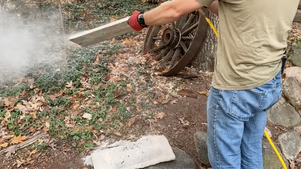 person in red gloves using a torch to clean metal chimney rods outdoors with leaves rocks and a tree nearby