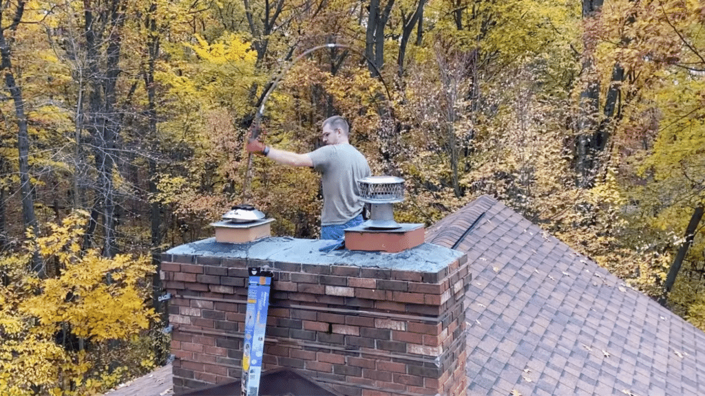 person on roof cleaning chimney with flexible rod near brick chimney and metal caps with fall trees behind