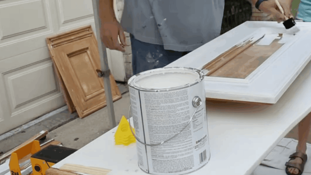 person painting wooden cabinet door with white paint using brush on table beside open paint can outside garage