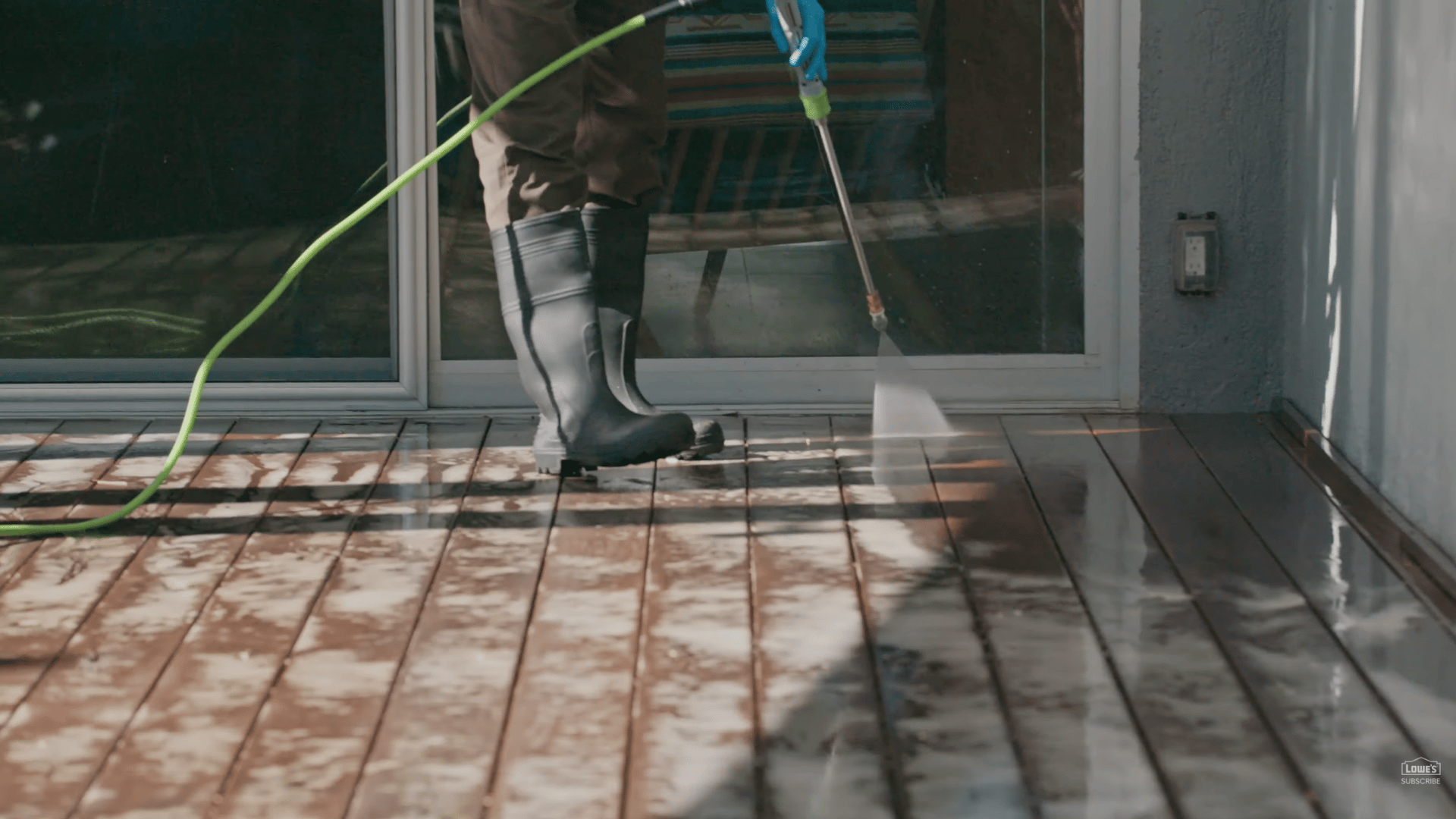 person pressure washing wooden deck near glass doors while wearing black boots and using green hose