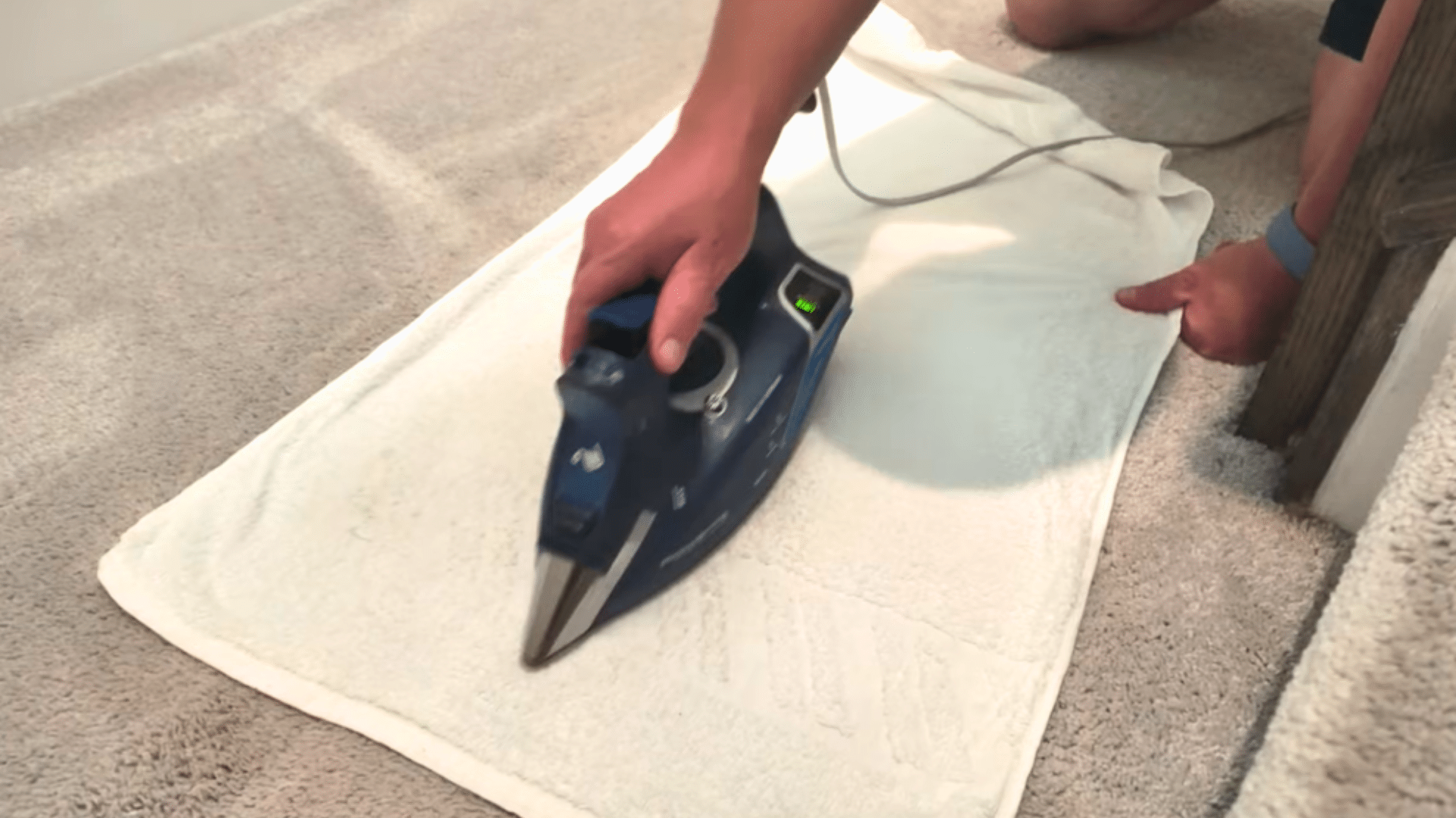 person repeating the steaming step with an iron over a white towel to loosen a tough carpet stain