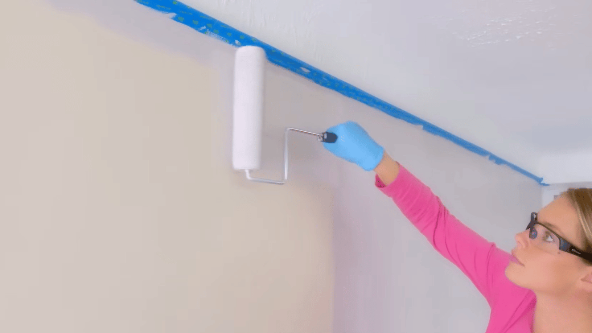 person rolling white paint along top edge of wall near ceiling with blue painter tape protecting trim line for clean finish
