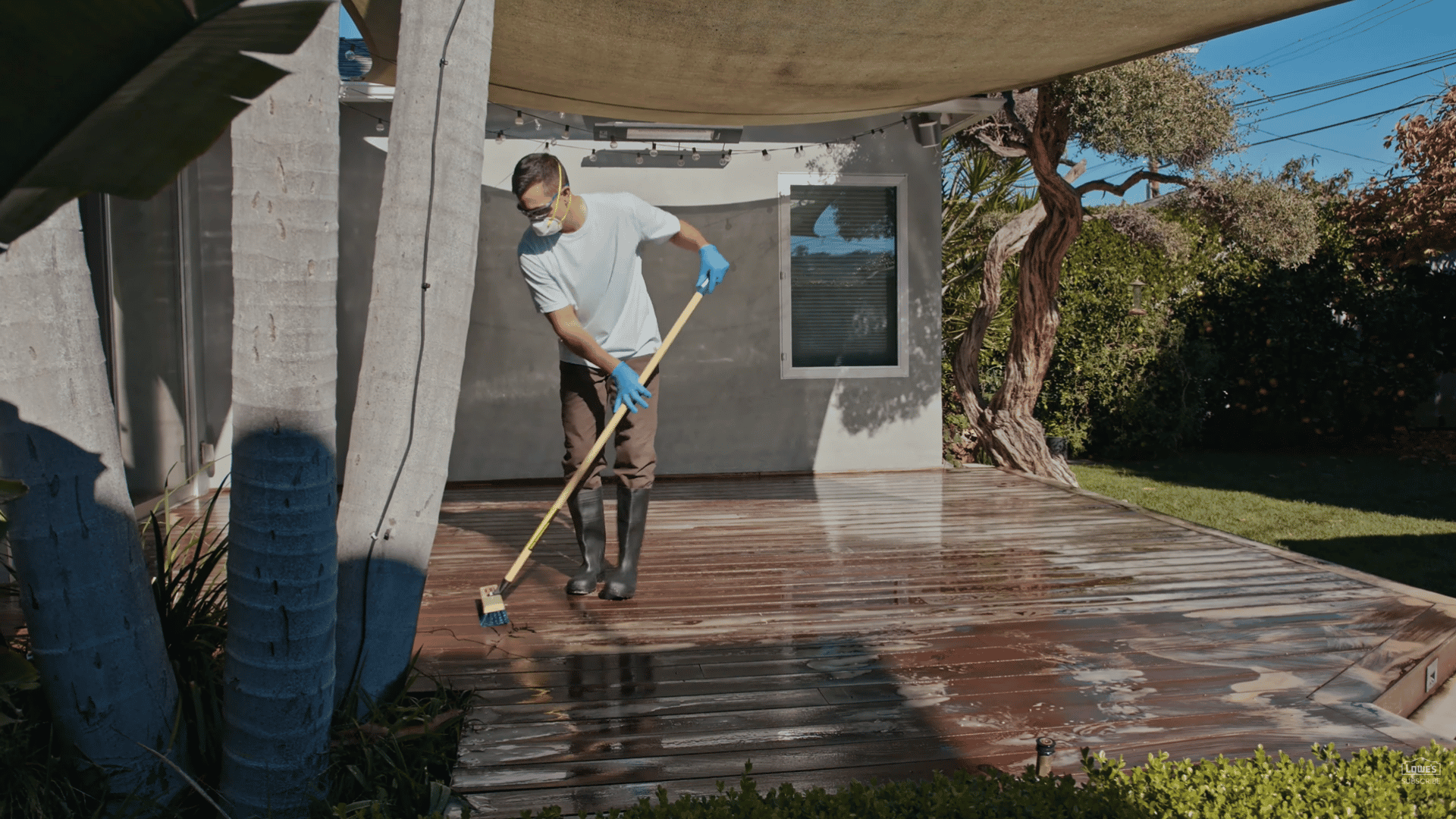 person scrubbing wet wooden patio deck while wearing gloves boots and face mask in sunny backyard