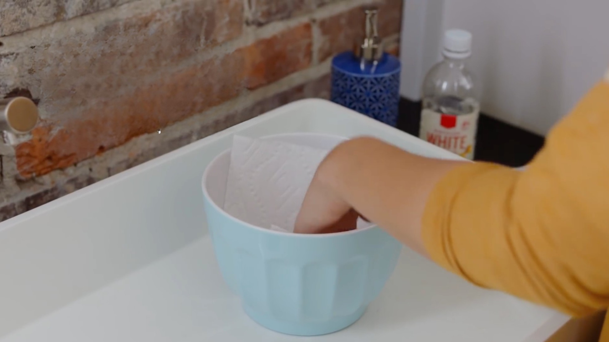 person soaks paper towels in vinegar inside a bowl to prepare for removing hard water stains from bathroom surfaces