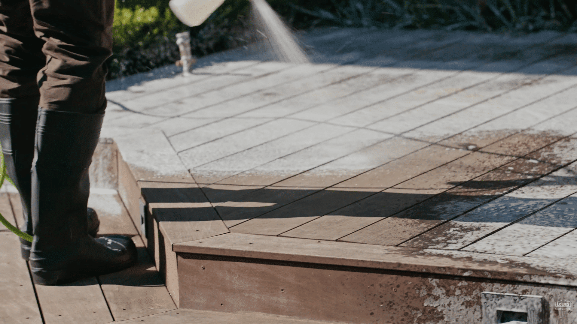 person spraying cleaner on outdoor wood deck while wearing black boots during patio maintenance