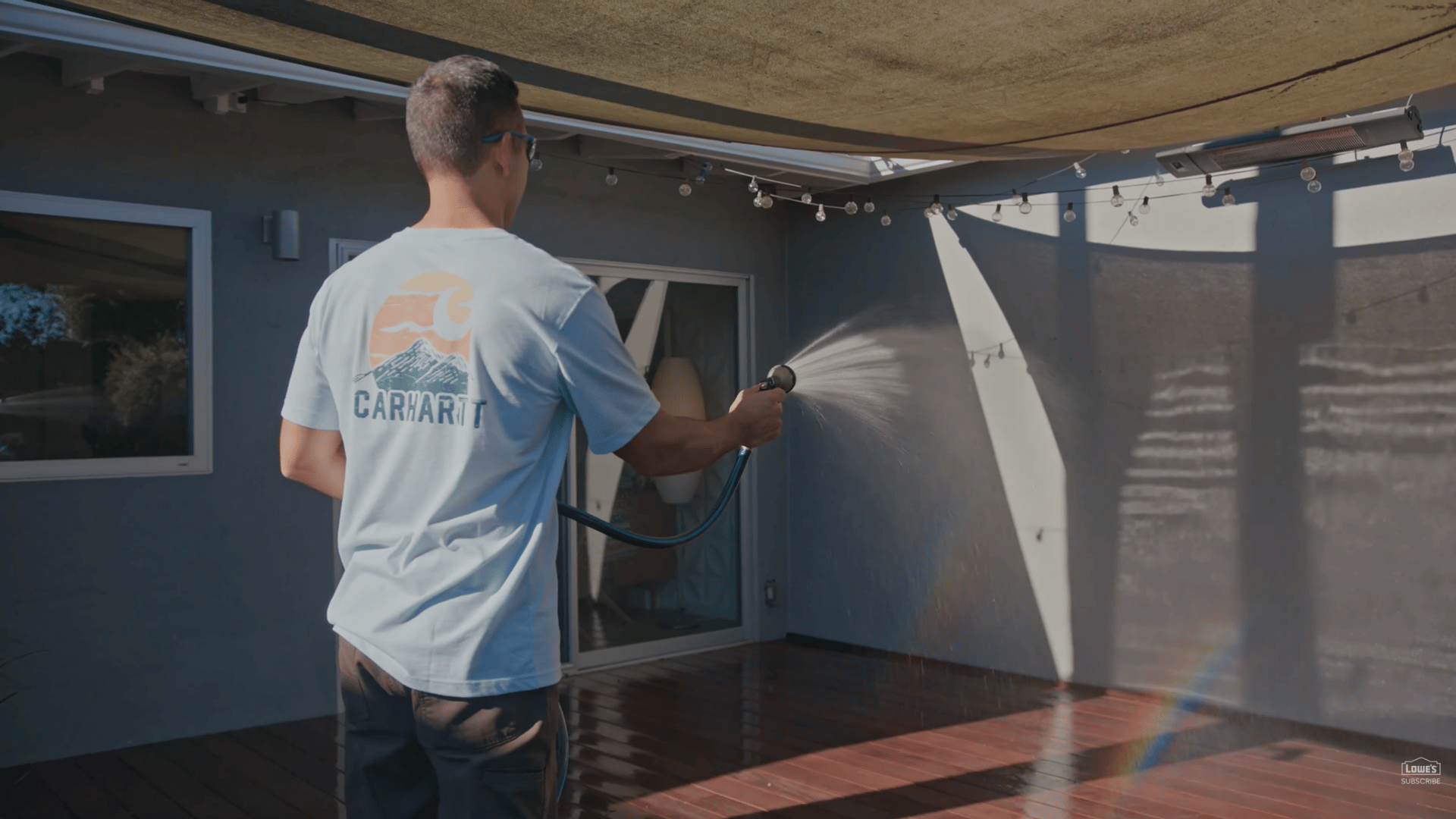 person spraying water across clean wooden patio deck under shade canopy in sunny backyard space