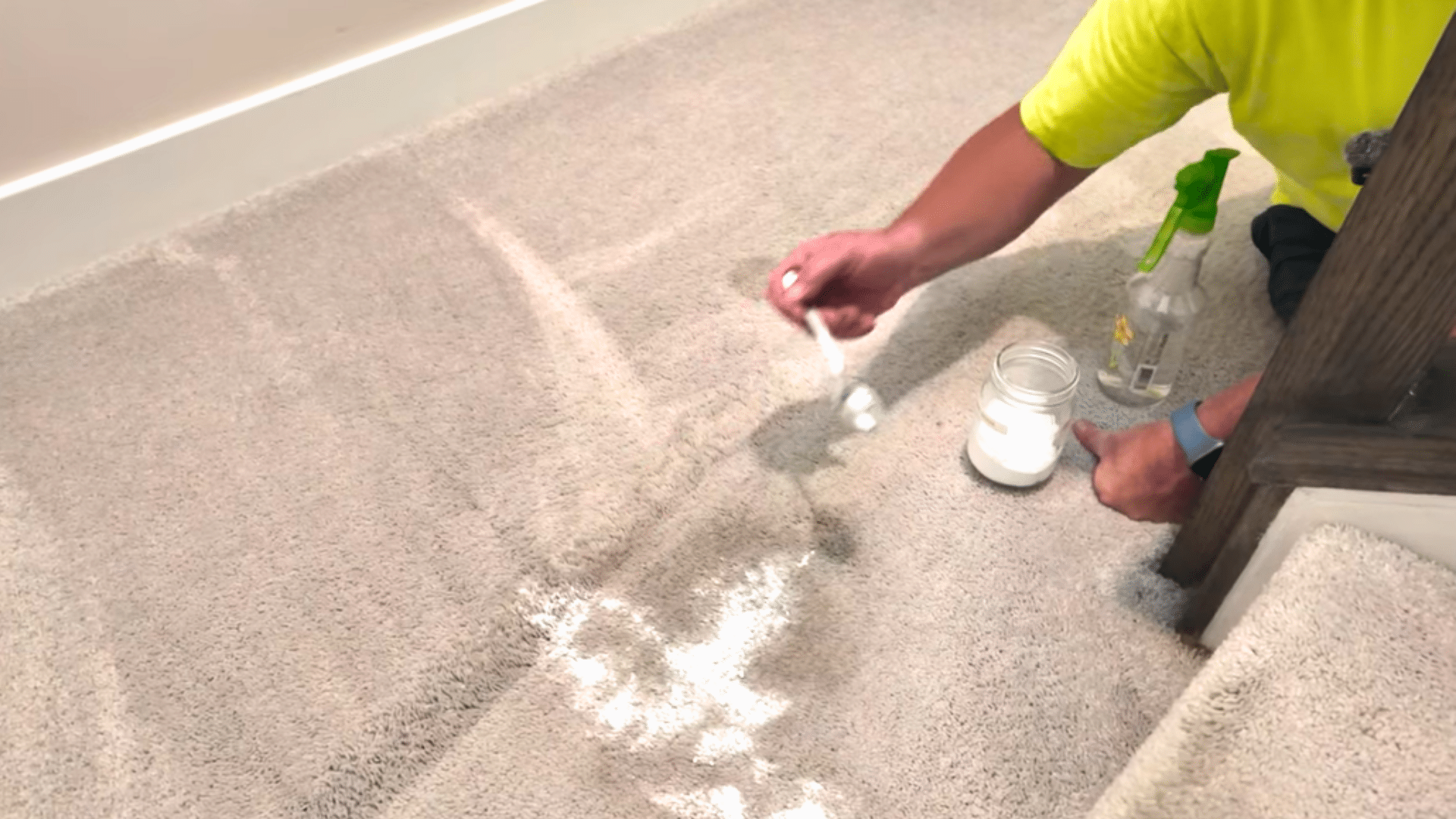 person sprinkling baking soda on a beige carpet beside a jar and spray bottle near a chair