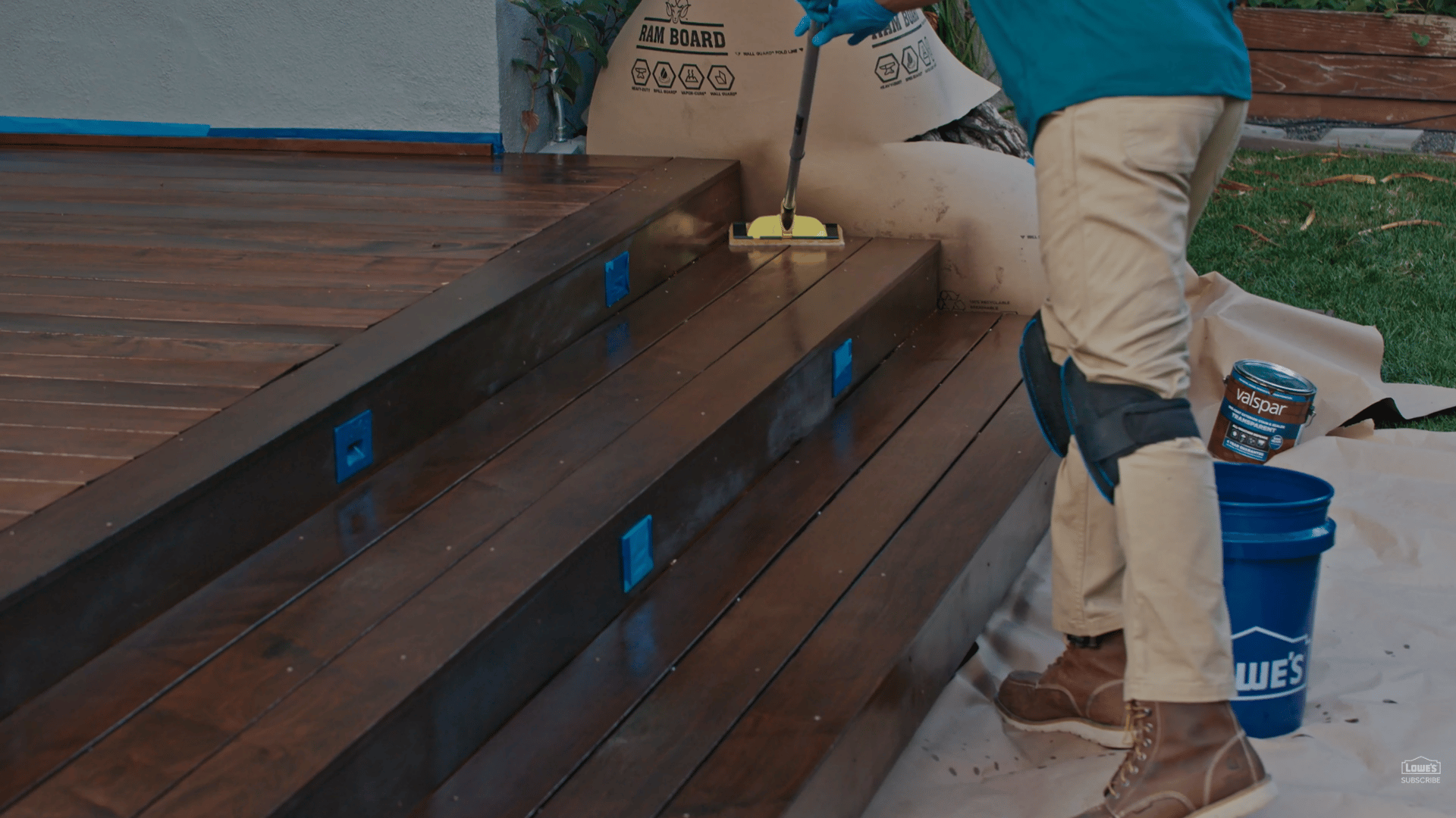 person staining dark wooden deck stairs with applicator pad beside blue bucket during refinishing project