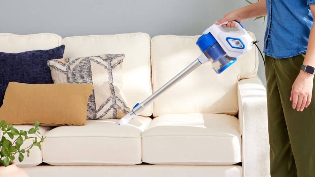 person using a handheld vacuum cleaner on a light colored sofa with decorative pillows in a bright living room setting
