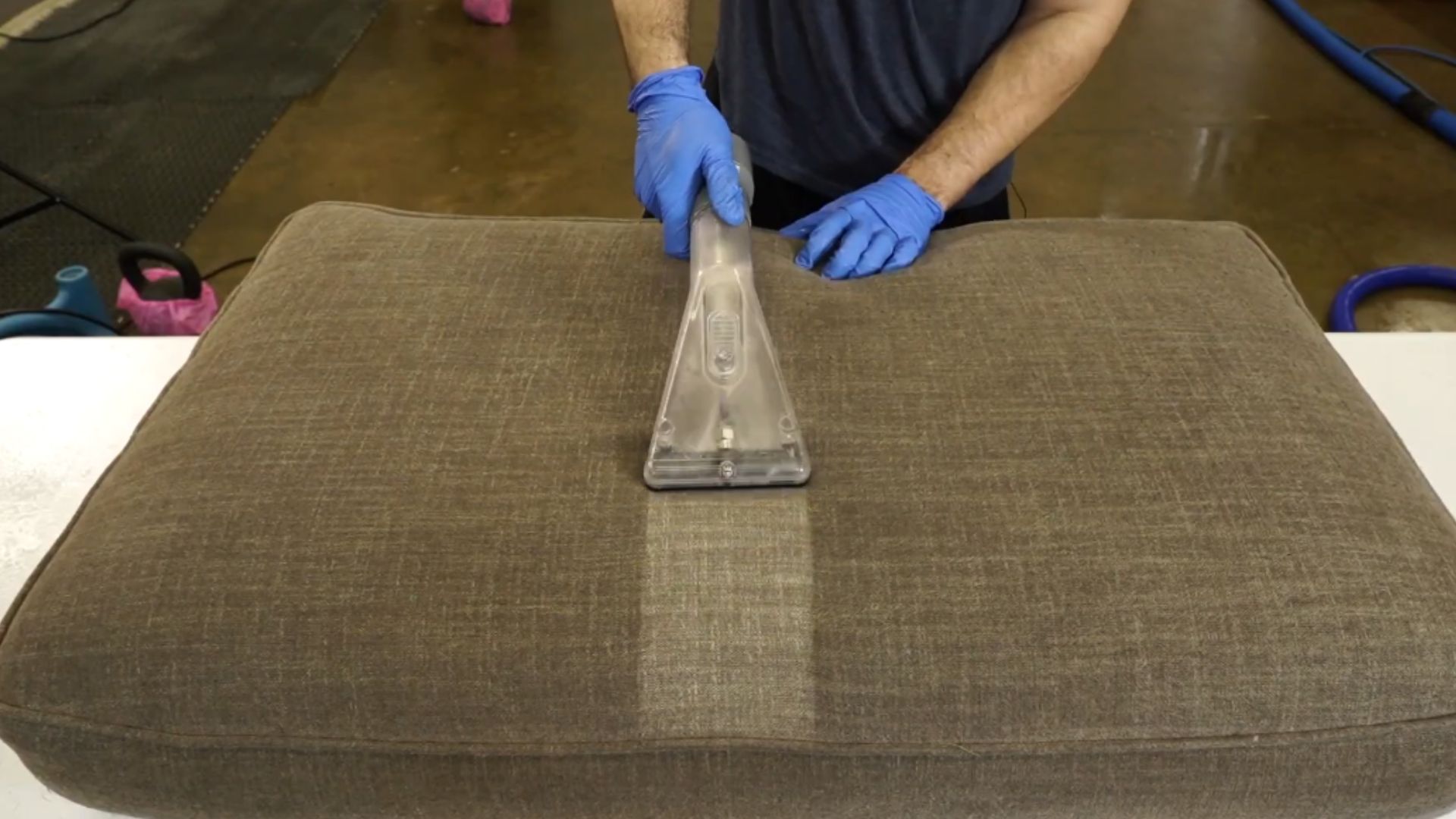 person wearing blue gloves using upholstery extraction tool to clean large fabric cushion removing dirt and stains