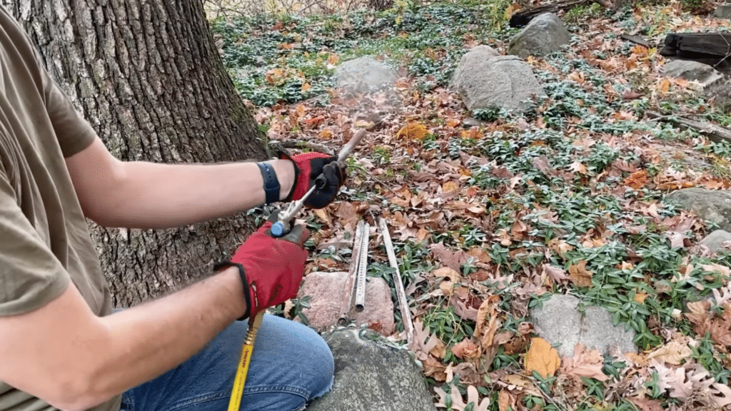 person wearing red gloves uses a torch to clean metal chimney rods outdoors with leaves rocks and a tree visible nearby