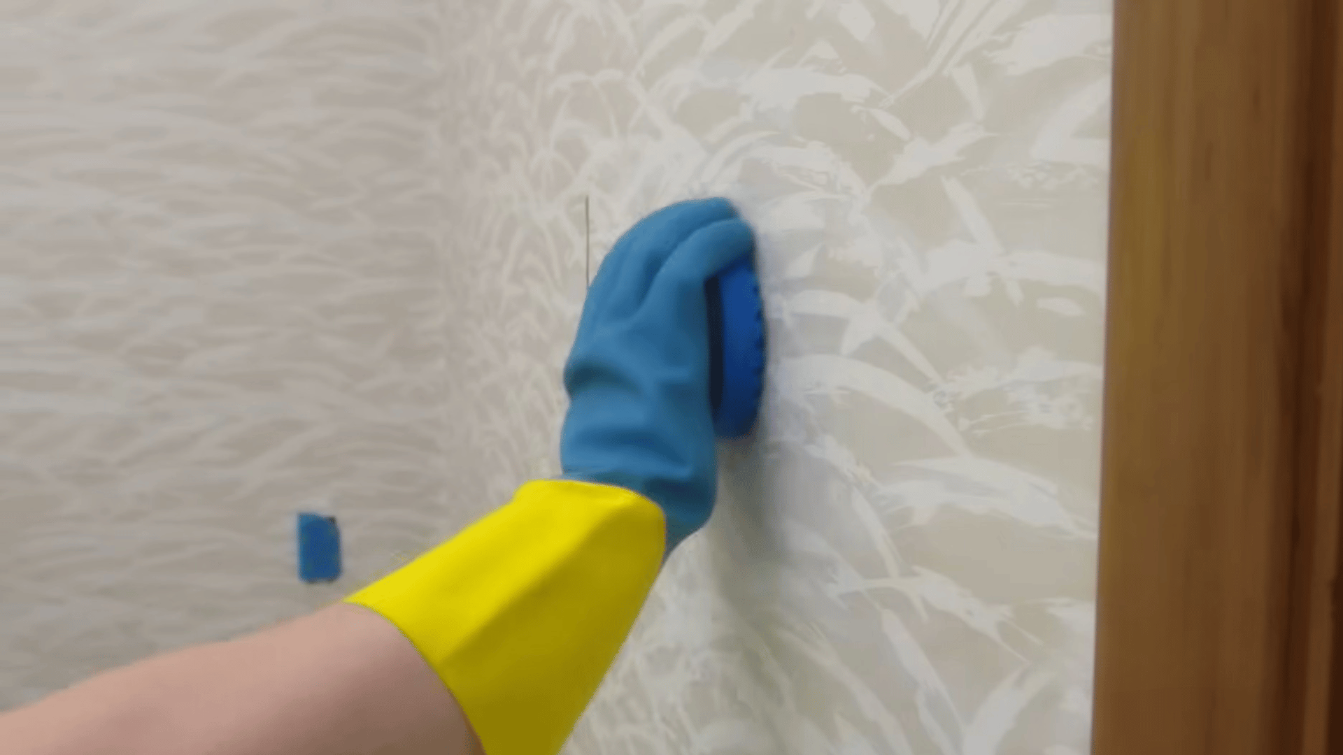 person wearing yellow and blue gloves scrubbing soapy wall surface with sponge during cleaning or wall preparation before painting