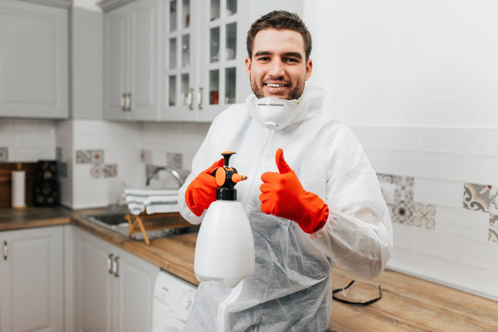 Man in protective suit with spray bottle giving thumbs up in a modern kitchen