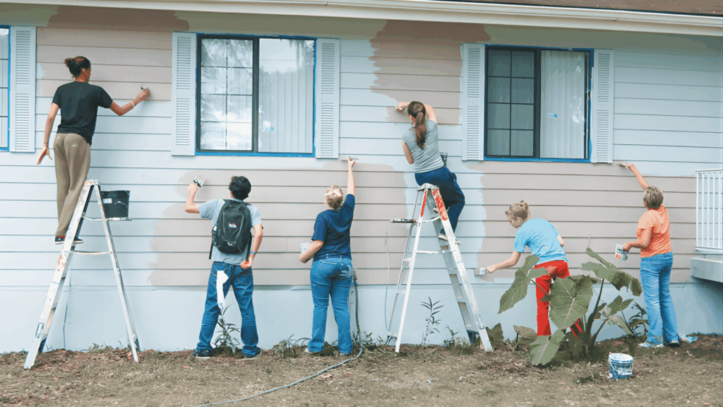 six people on ladders painting exterior wall of a house together in home renovation to reduce how much it costs to paint house