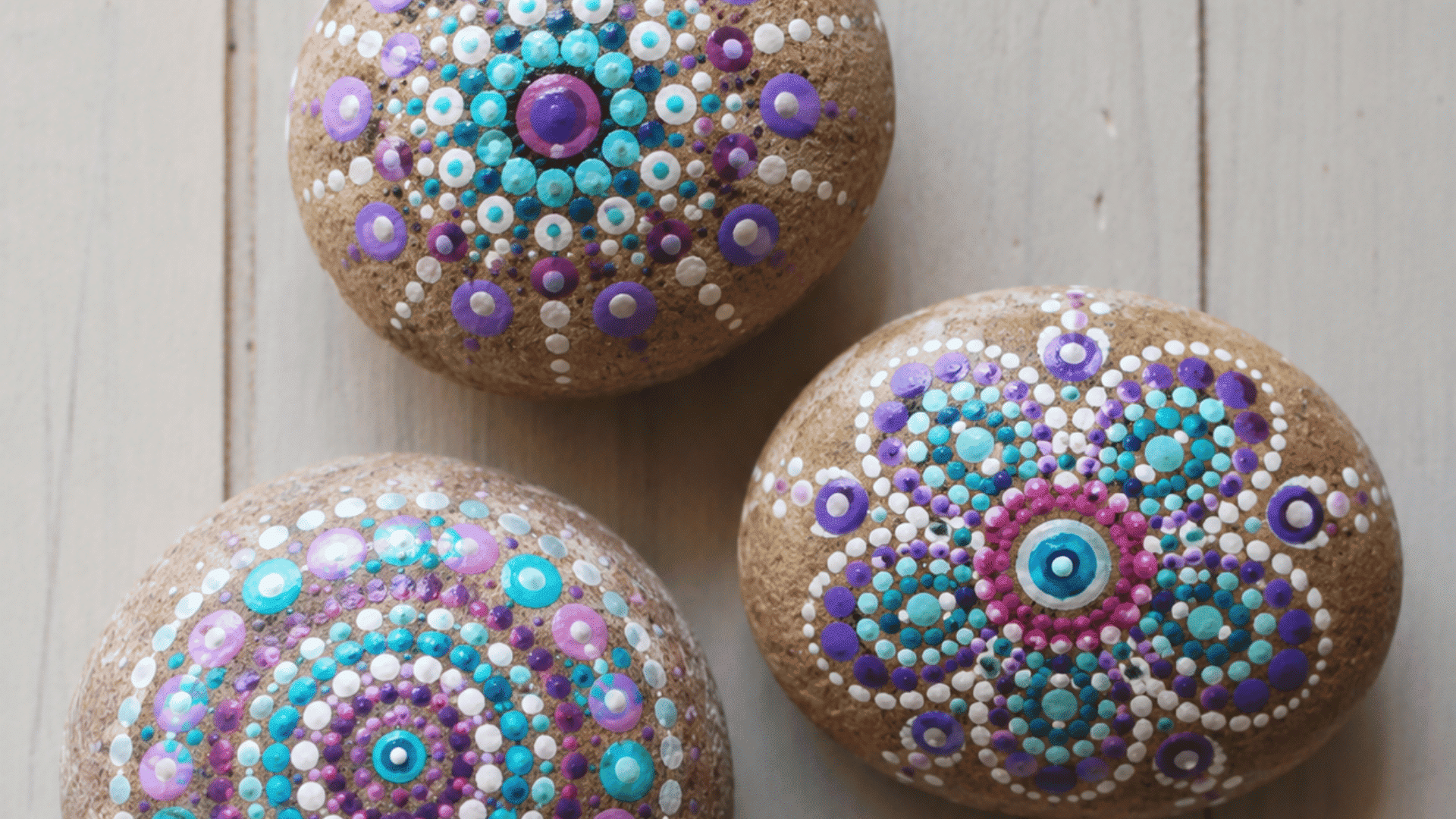 three smooth rocks with colorful dot mandala patterns placed on light wooden surface in natural lighting