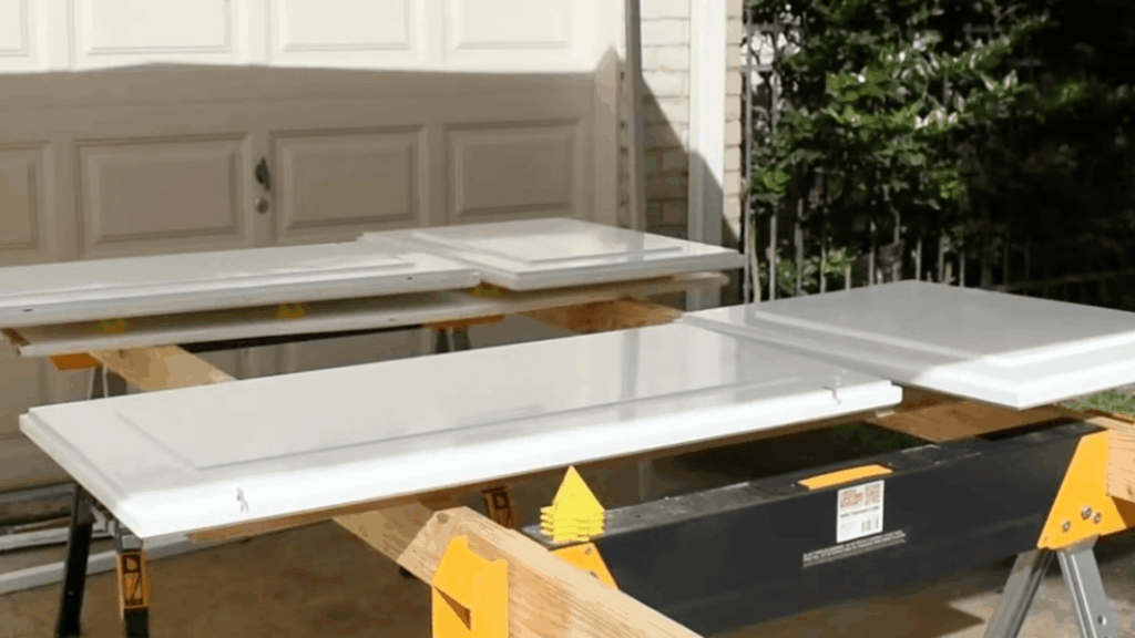 three white painted cabinet doors drying on sawhorses outdoors in front of garage with sunlight and greenery nearby