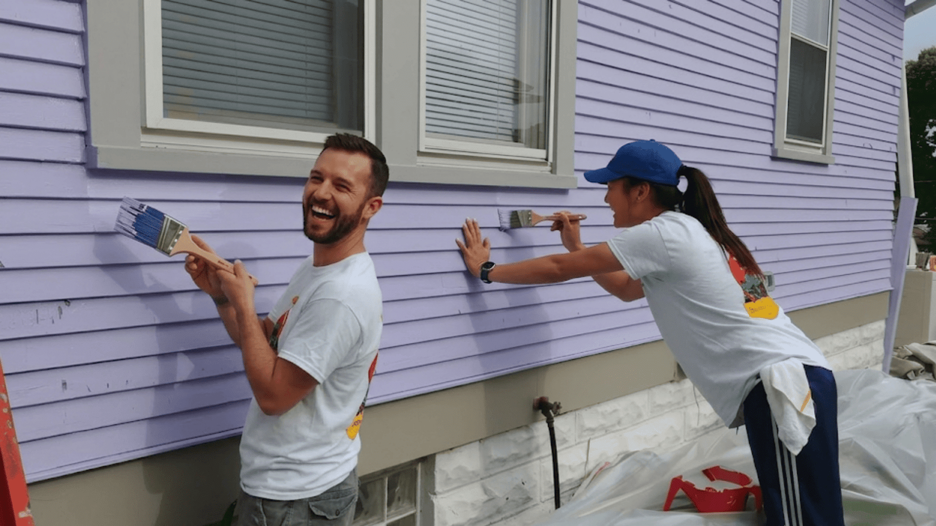 two people smiling and painting white wooden house exterior wall with brushes during diy project