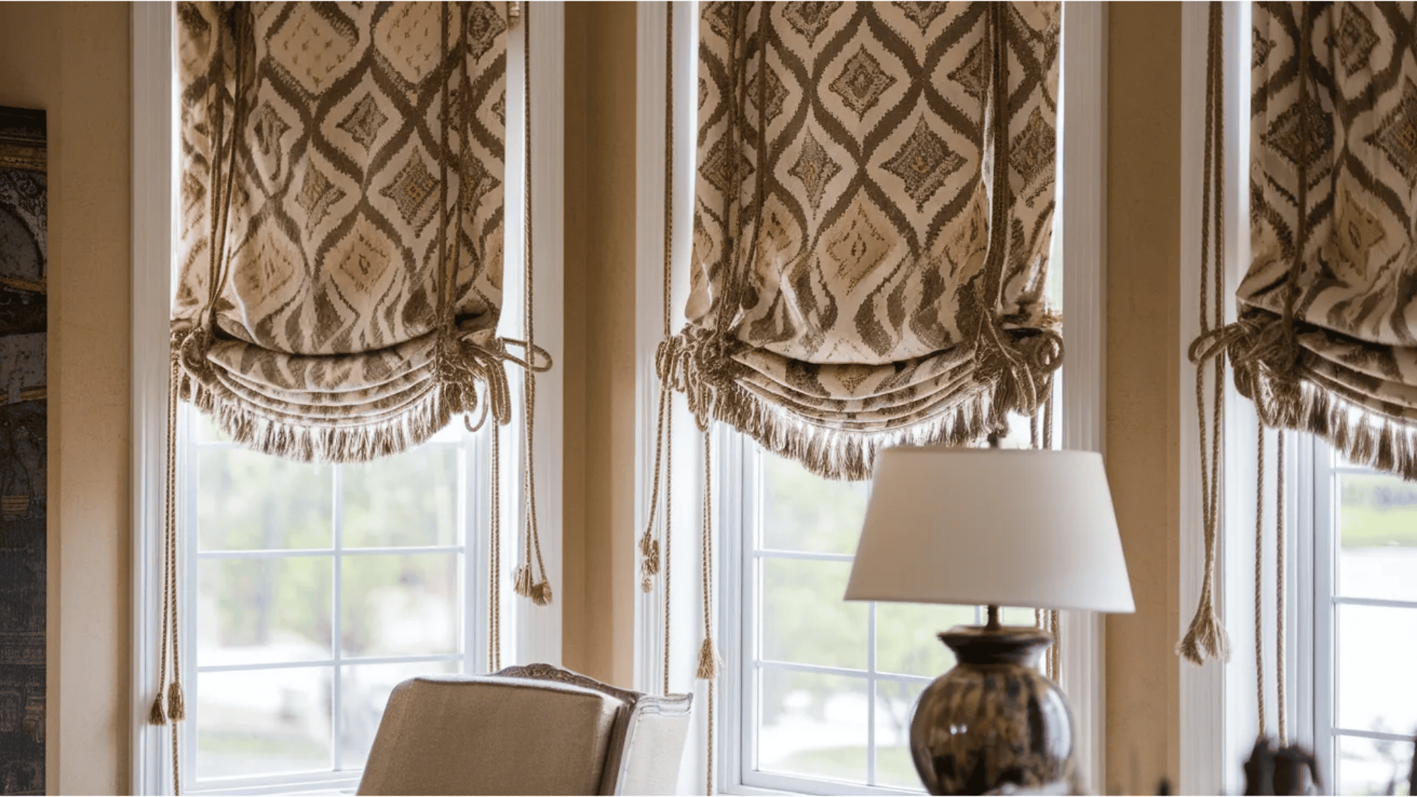 what is a roman shade patterned roman shade with tassels hanging over tall windows beside chair table and lamp