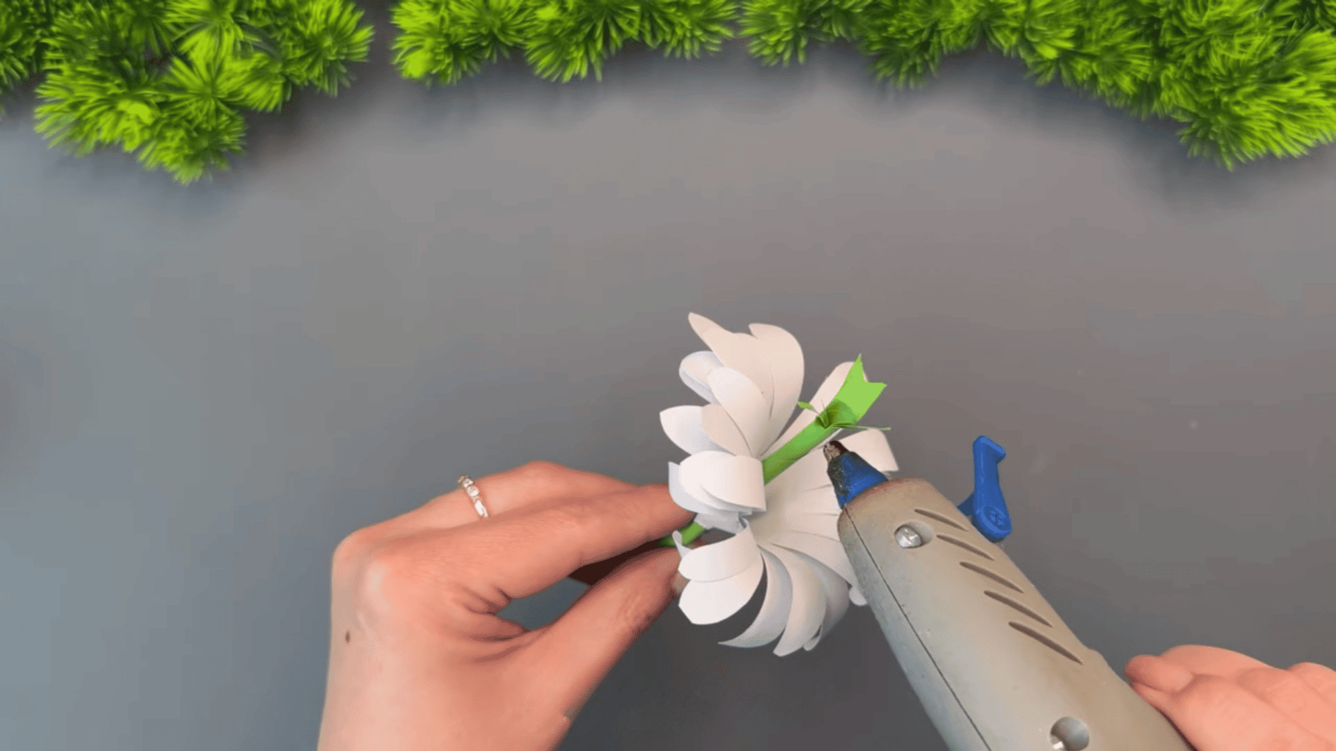 woman attaching curled white paper petals to a green paper stem with a hot glue gun while assembling a handmade flower