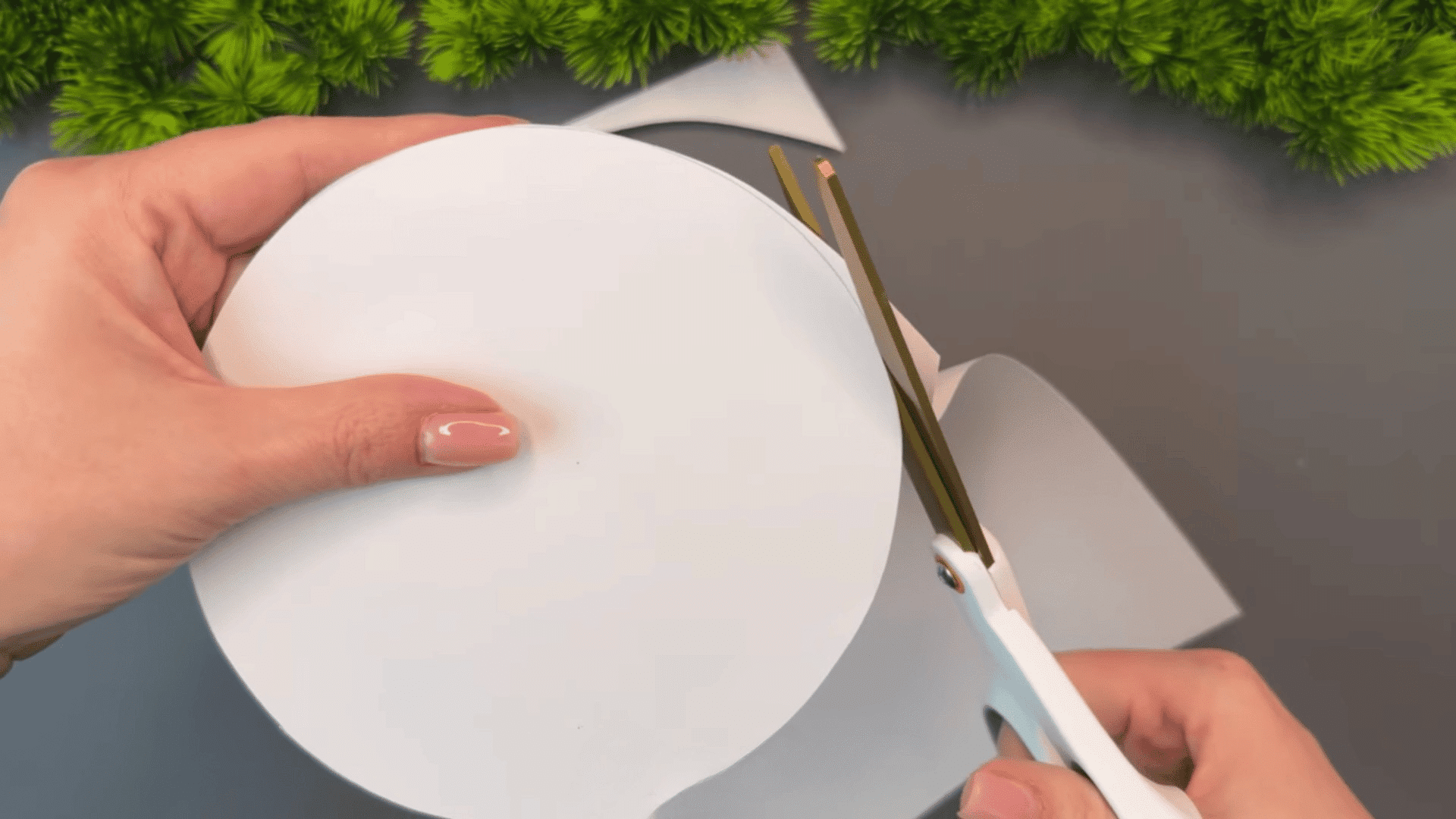 woman cutting a white paper circle with scissors on a gray table with green plants and paper scraps in the background