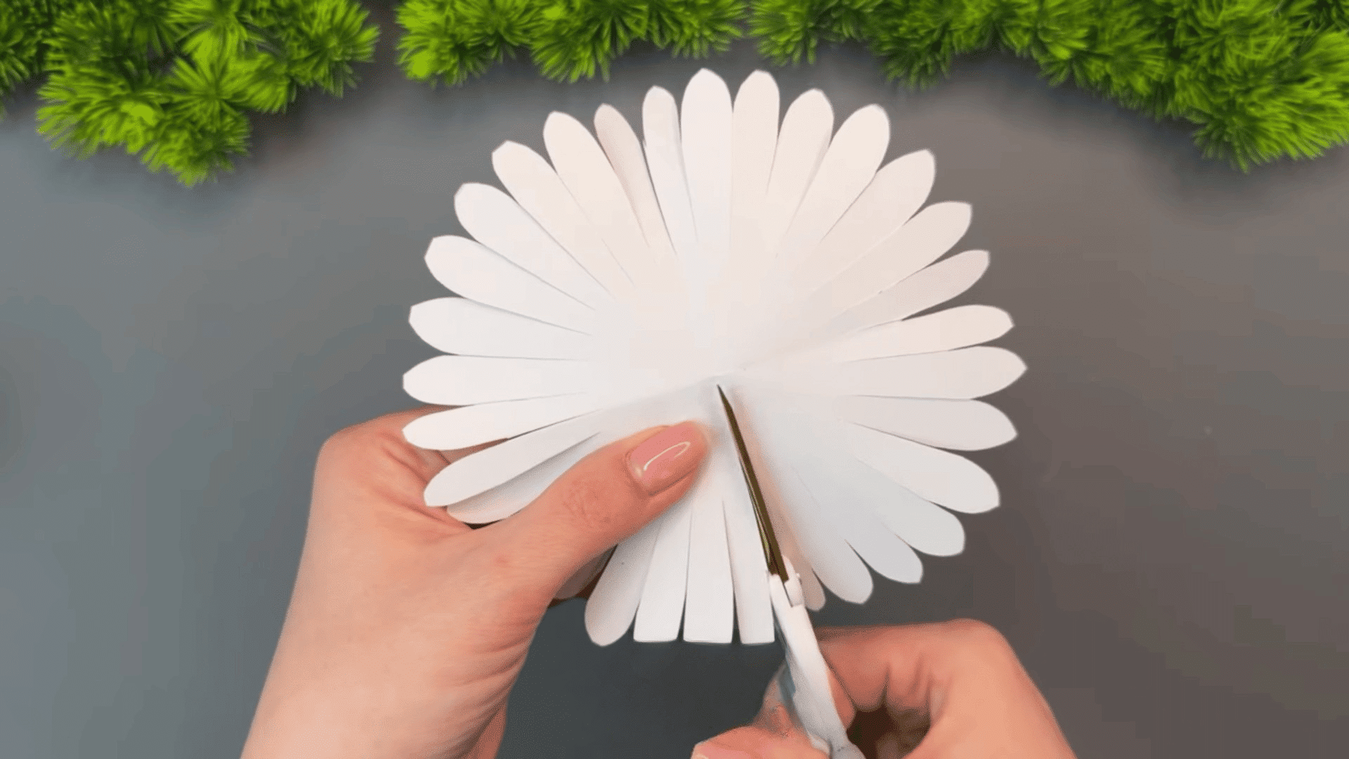 woman cutting the center of a white paper flower shape with scissors on a gray table decorated with green plants