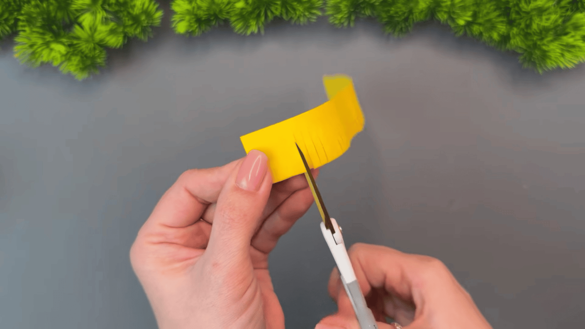 woman cutting thin fringe lines into folded yellow paper with scissors while making a paper flower craft on a gray table