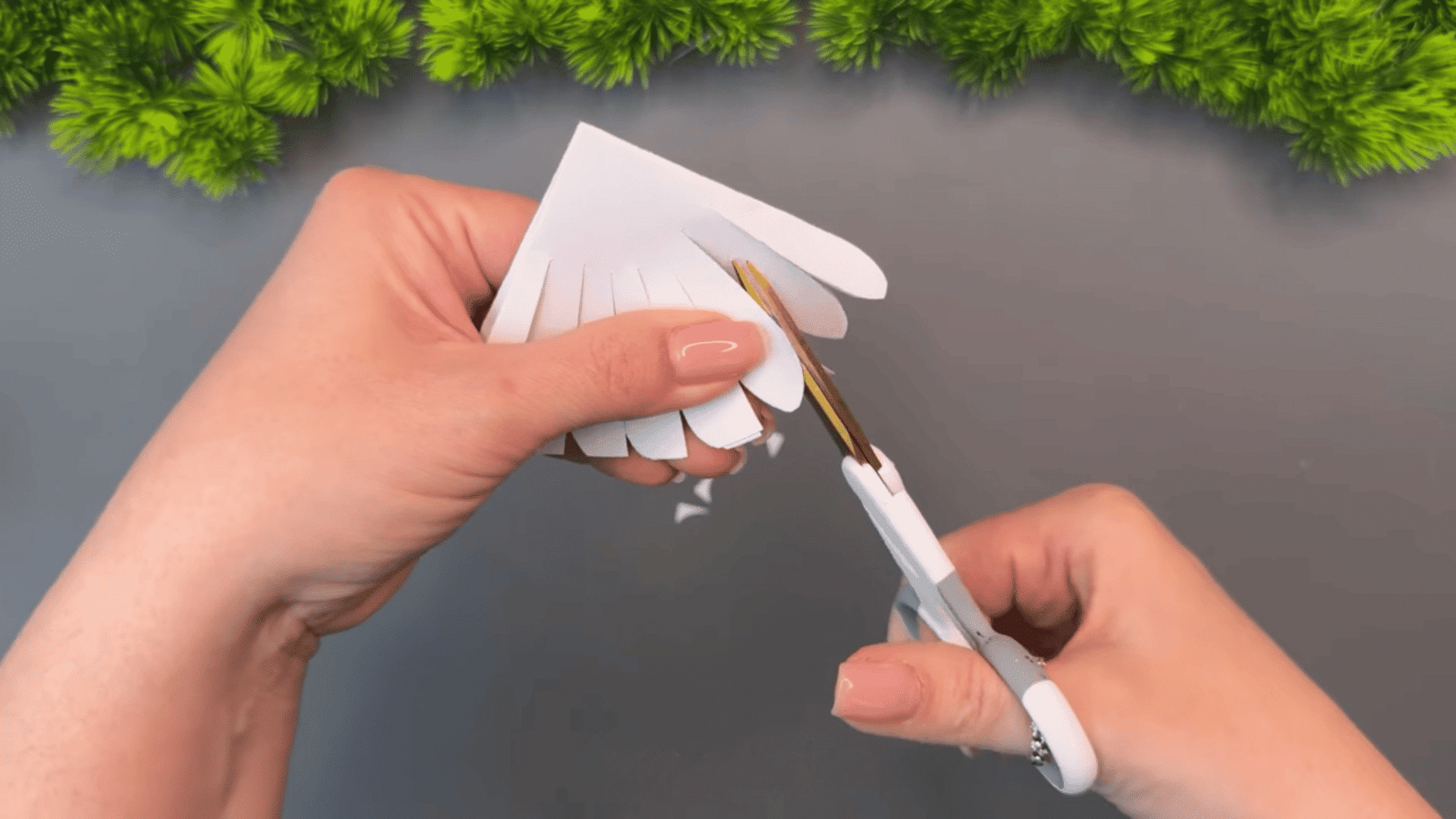 woman cutting thin strips into folded white paper with scissors while making a decorative craft on a gray table