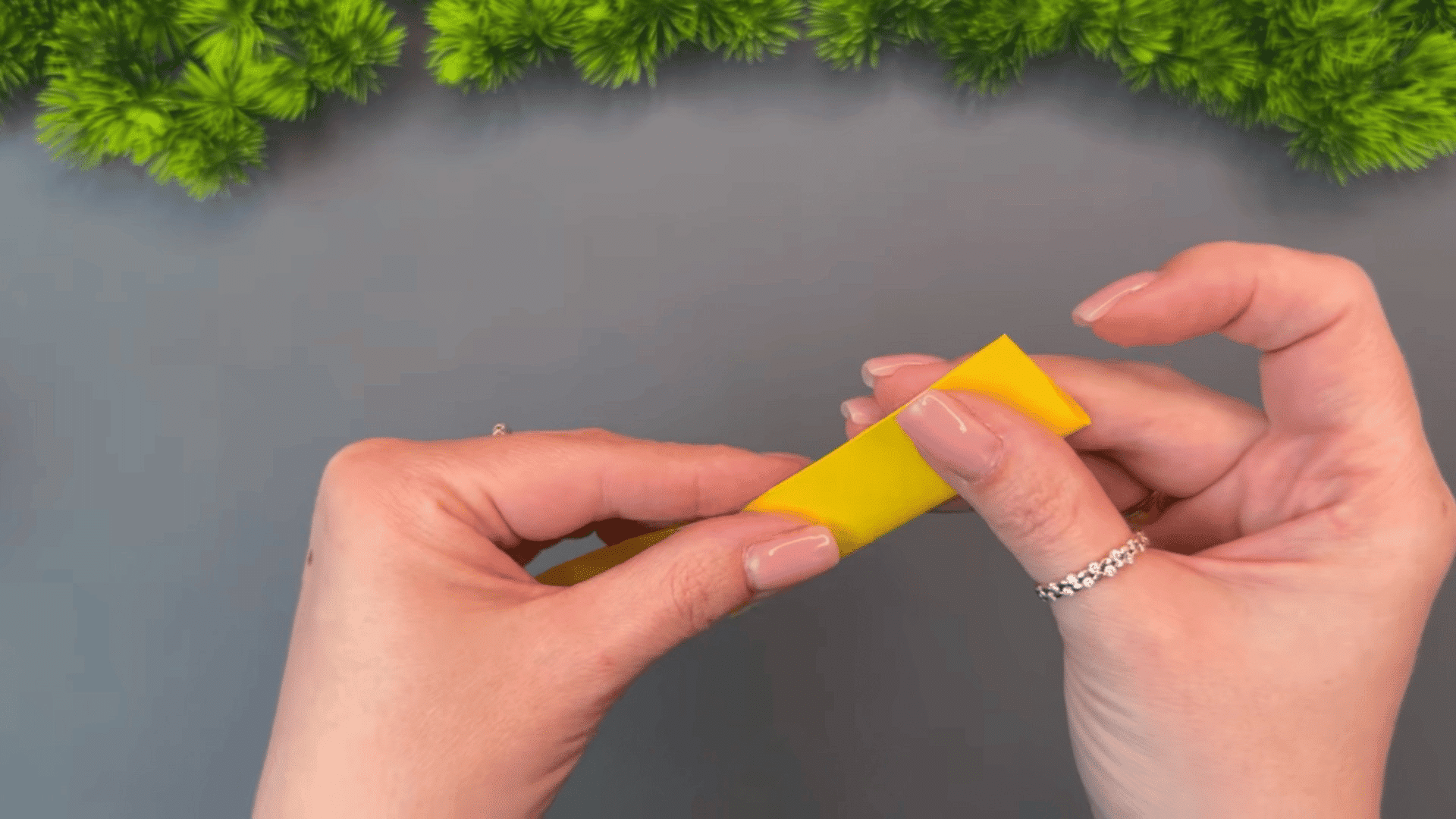 woman folding a bright yellow paper strip into a compact layered shape while crafting on a gray table with green plants