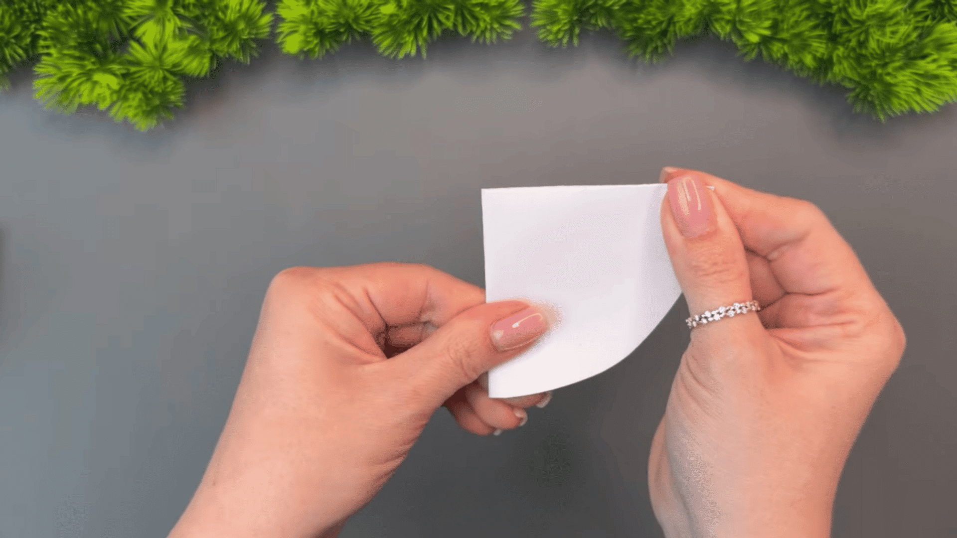 woman folding a white paper circle in half with both hands on a gray table decorated with green artificial plants