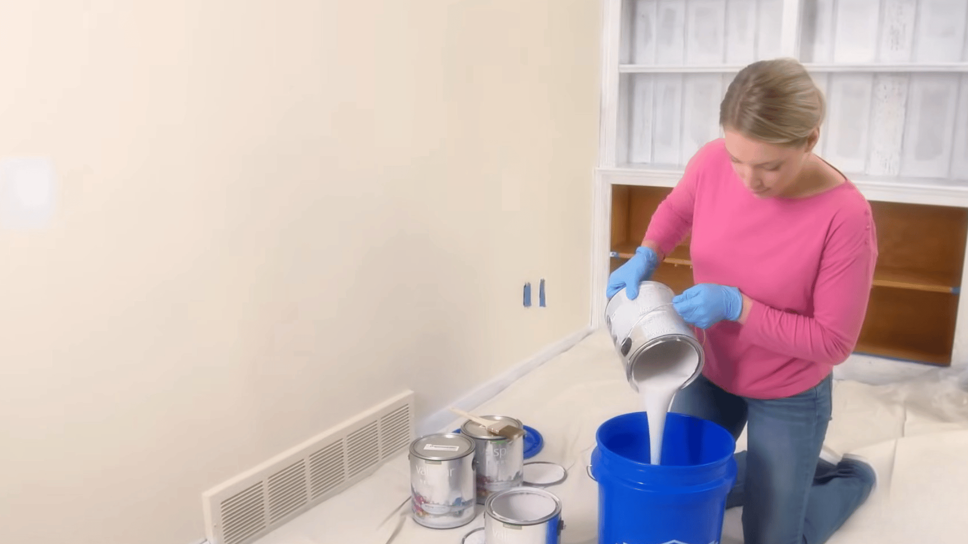 woman kneeling on floor pouring white paint from can into blue bucket with several paint cans nearby in a room being prepared