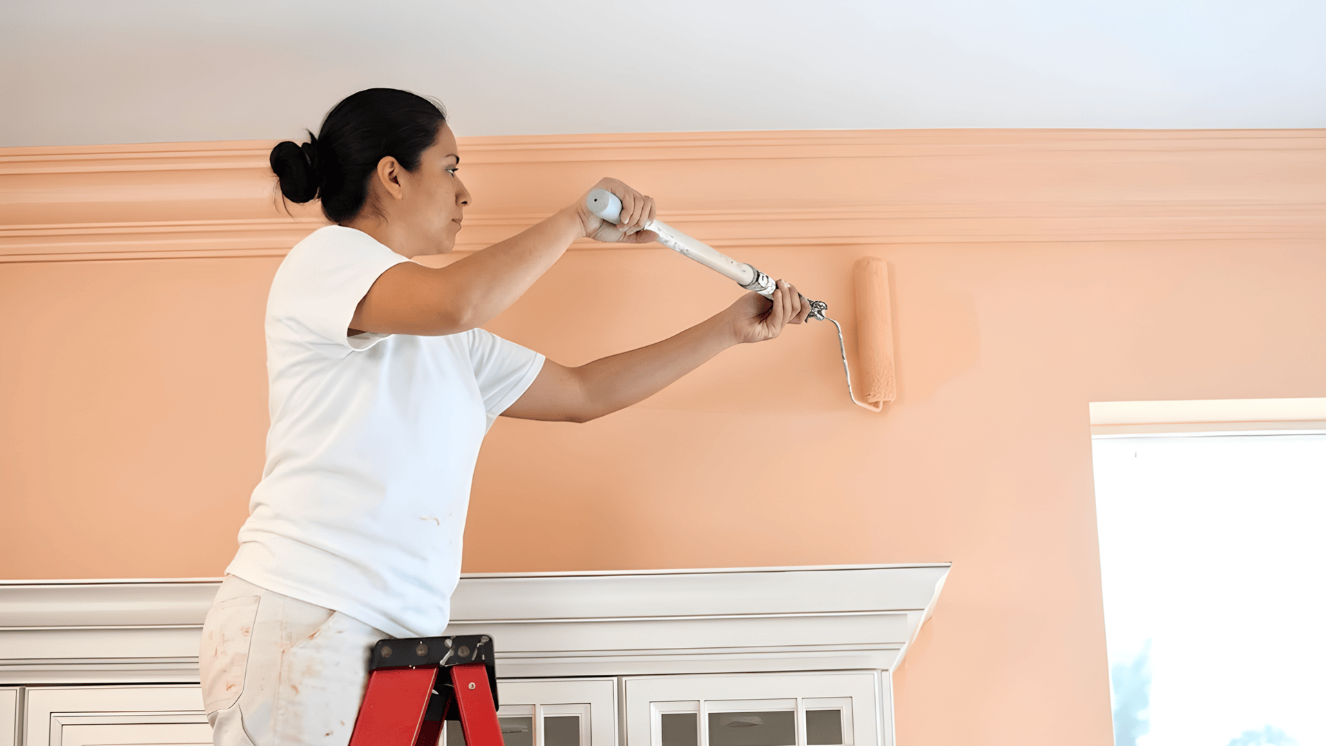 woman on ladder painting interior wall with roller near ceiling trim during home improvement project