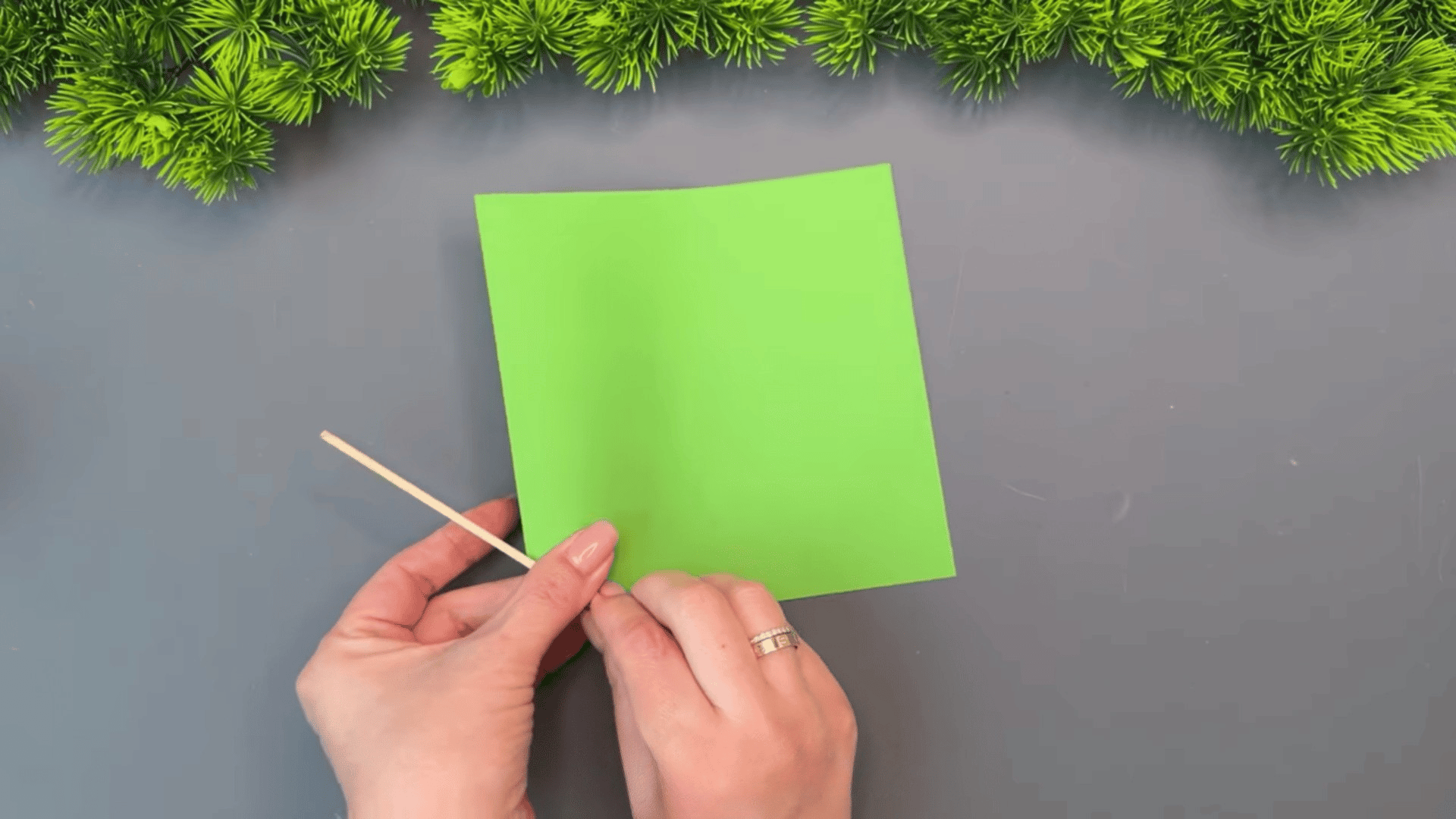 woman placing a wooden stick beside a bright green paper sheet on a gray table while preparing craft materials