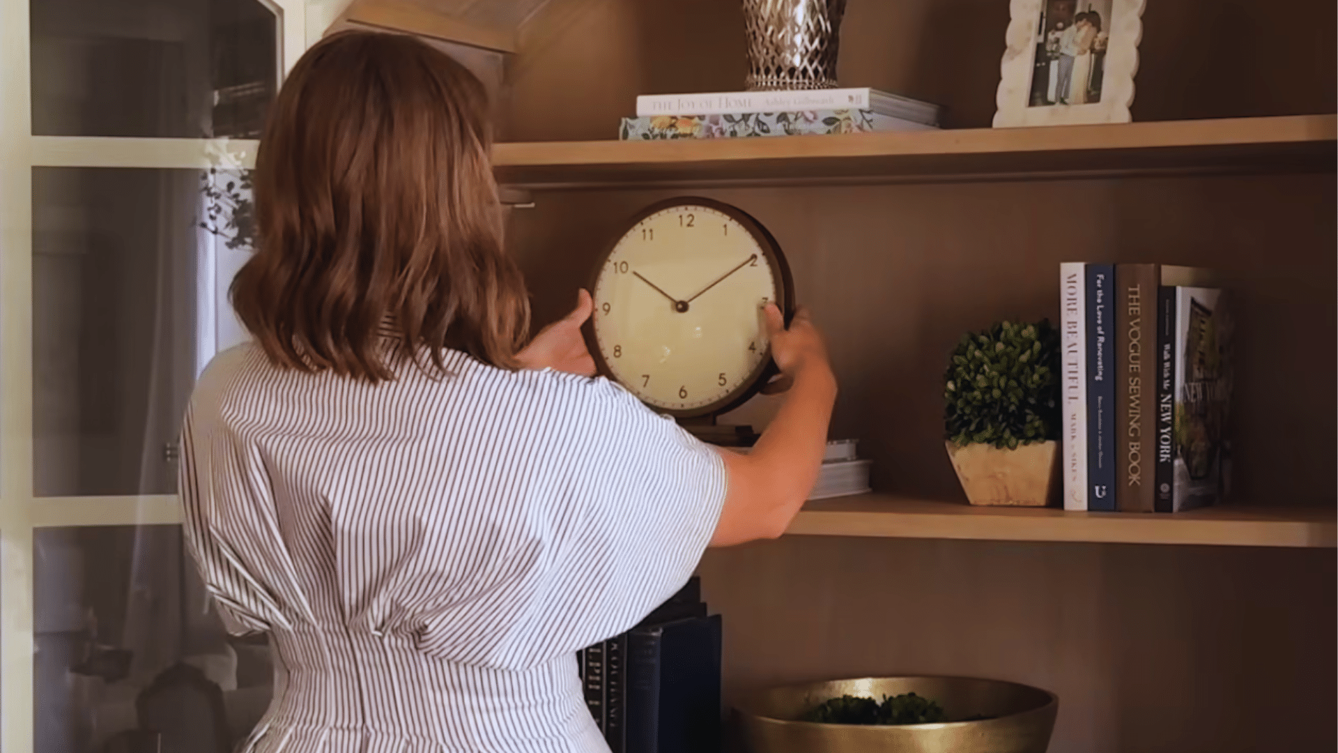woman placing round clock on shelf balancing tall and short items among books and decor in bookshelf