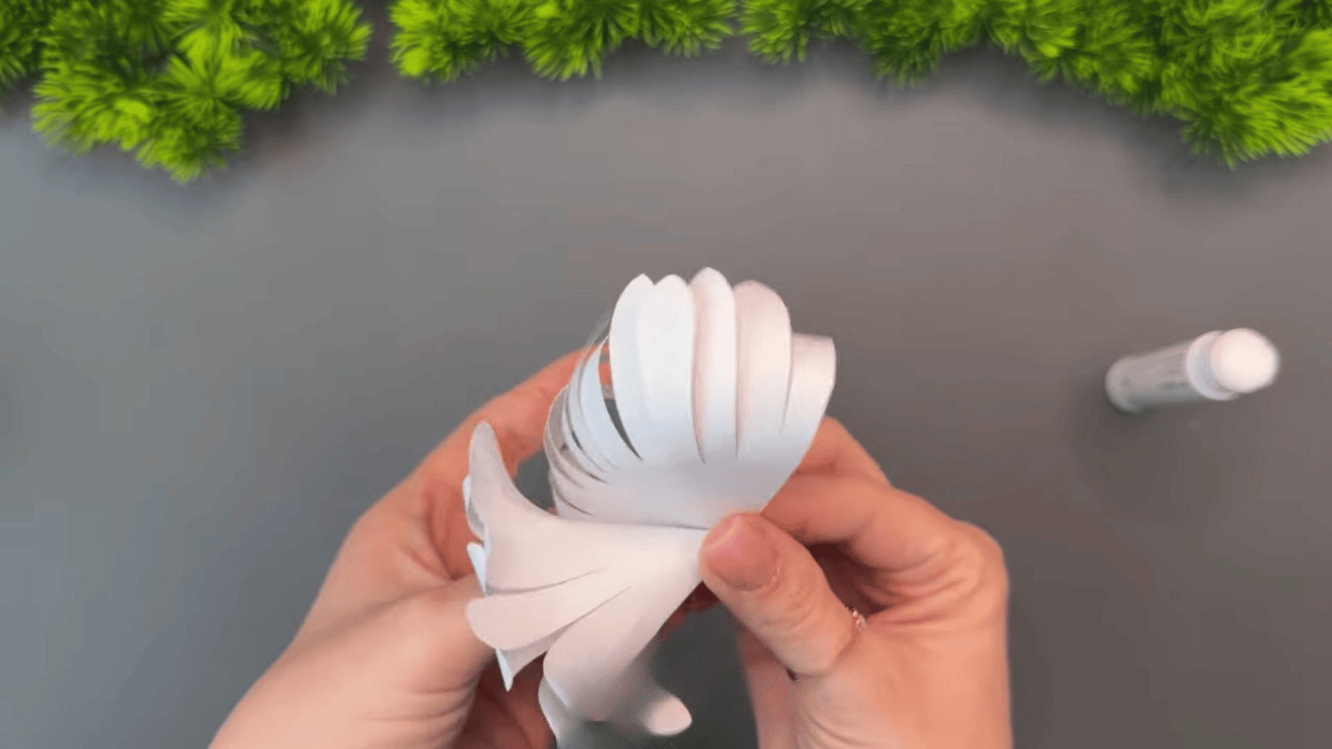 woman rolling and gluing a white paper flower shape into a layered cone design on a gray table with green decorations