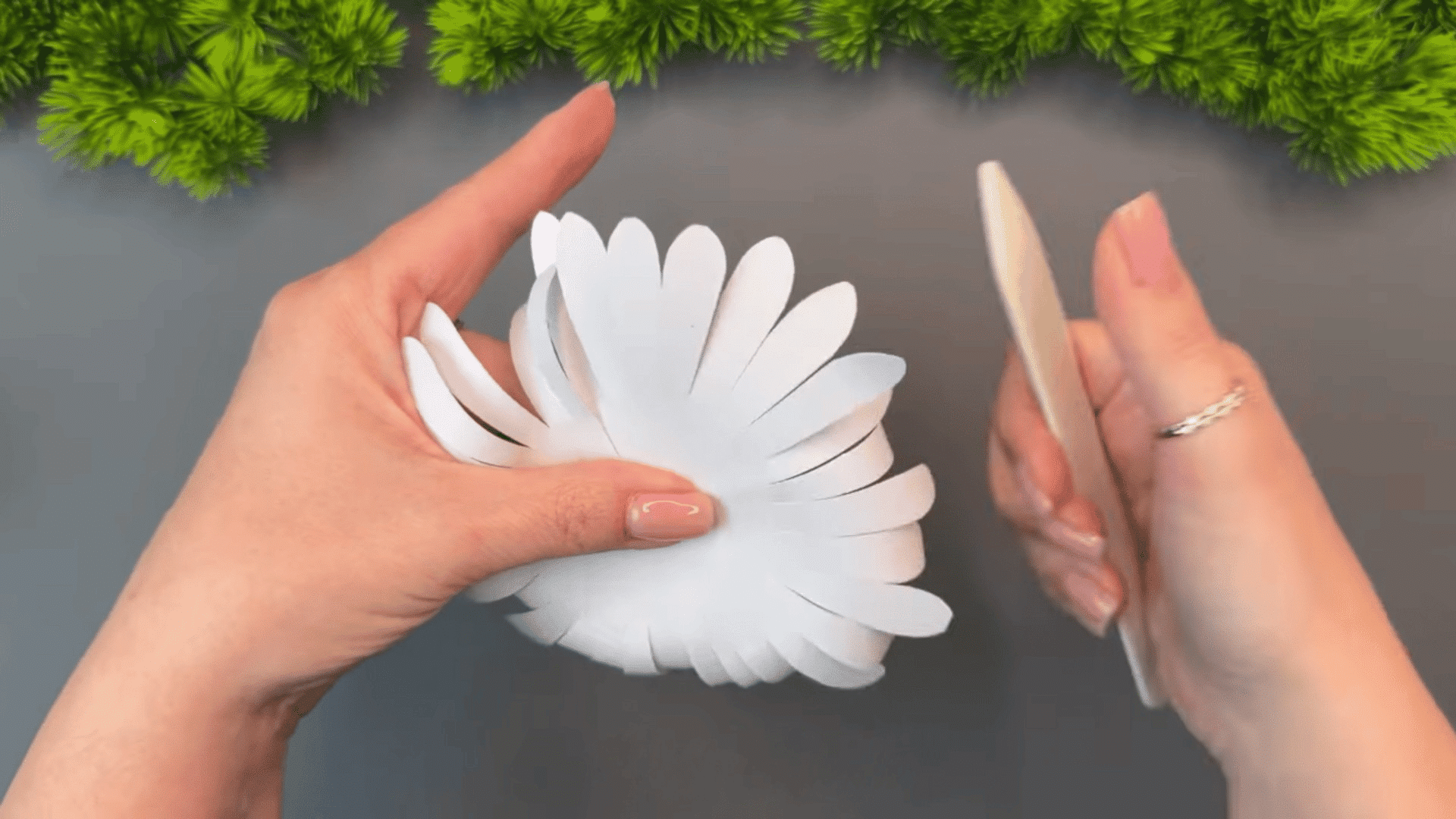 woman shaping curled white paper petals with a wooden stick while holding a handmade paper flower on a gray table