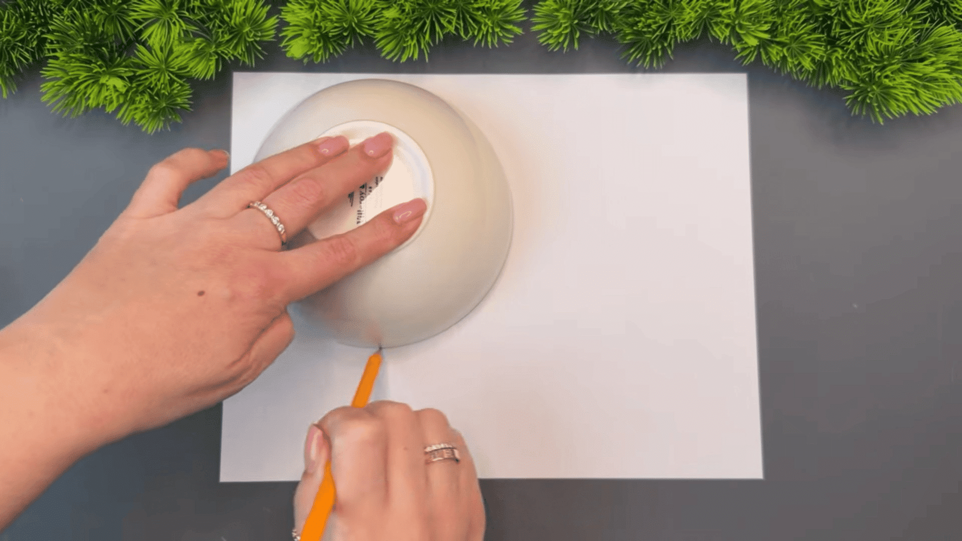woman tracing a circle on white paper with a pencil using an upside down bowl on a gray table with green plants along the edge