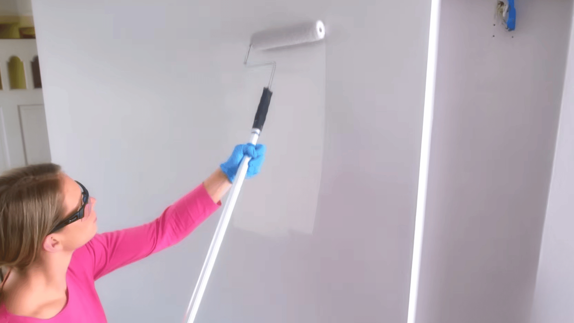 woman using extended roller to paint upper section of tall wall while looking upward and maintaining even coat of paint