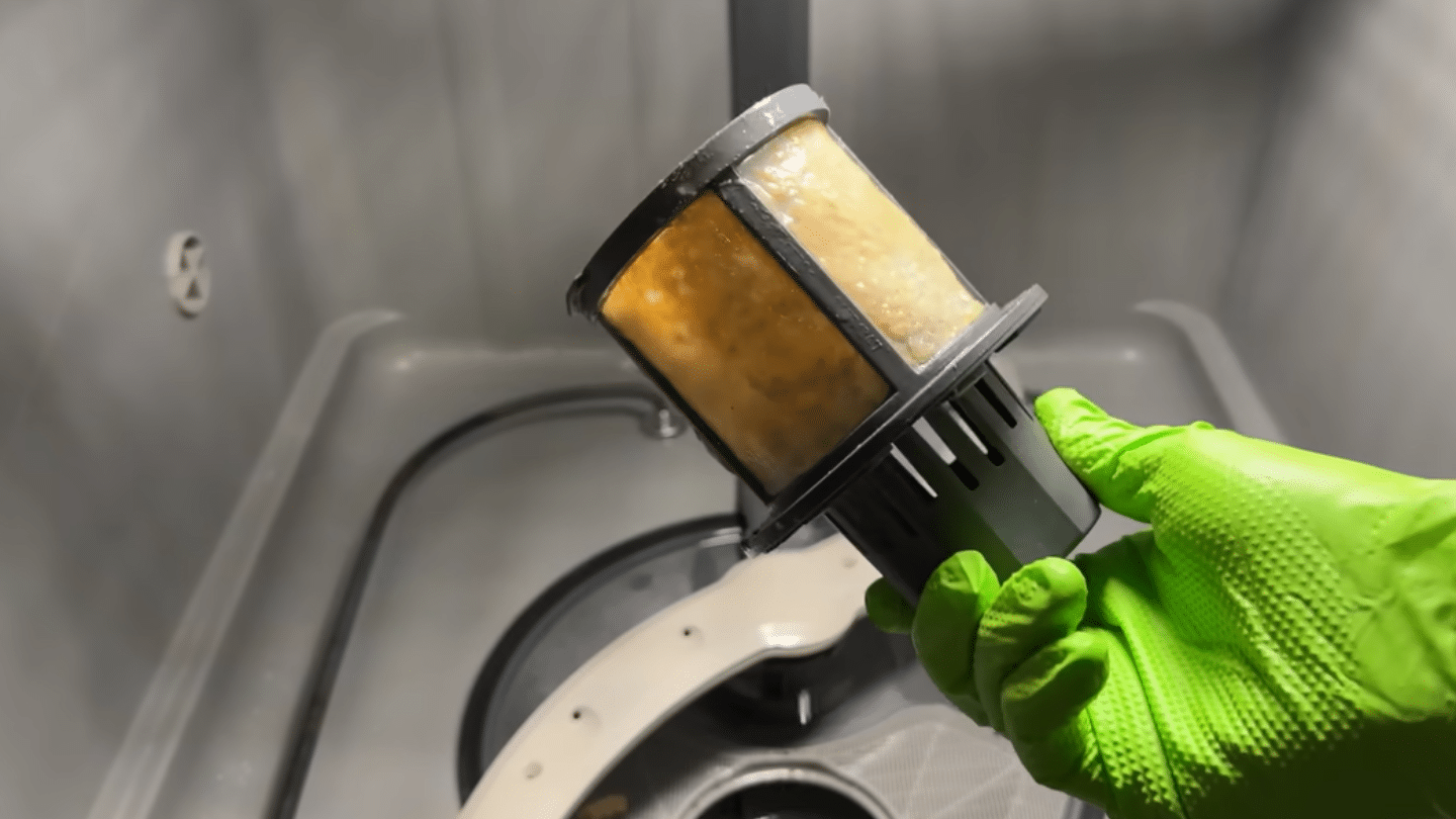 a close-up of a person in a bright green glove holding a dirty dishwasher filter covered in yellow grime