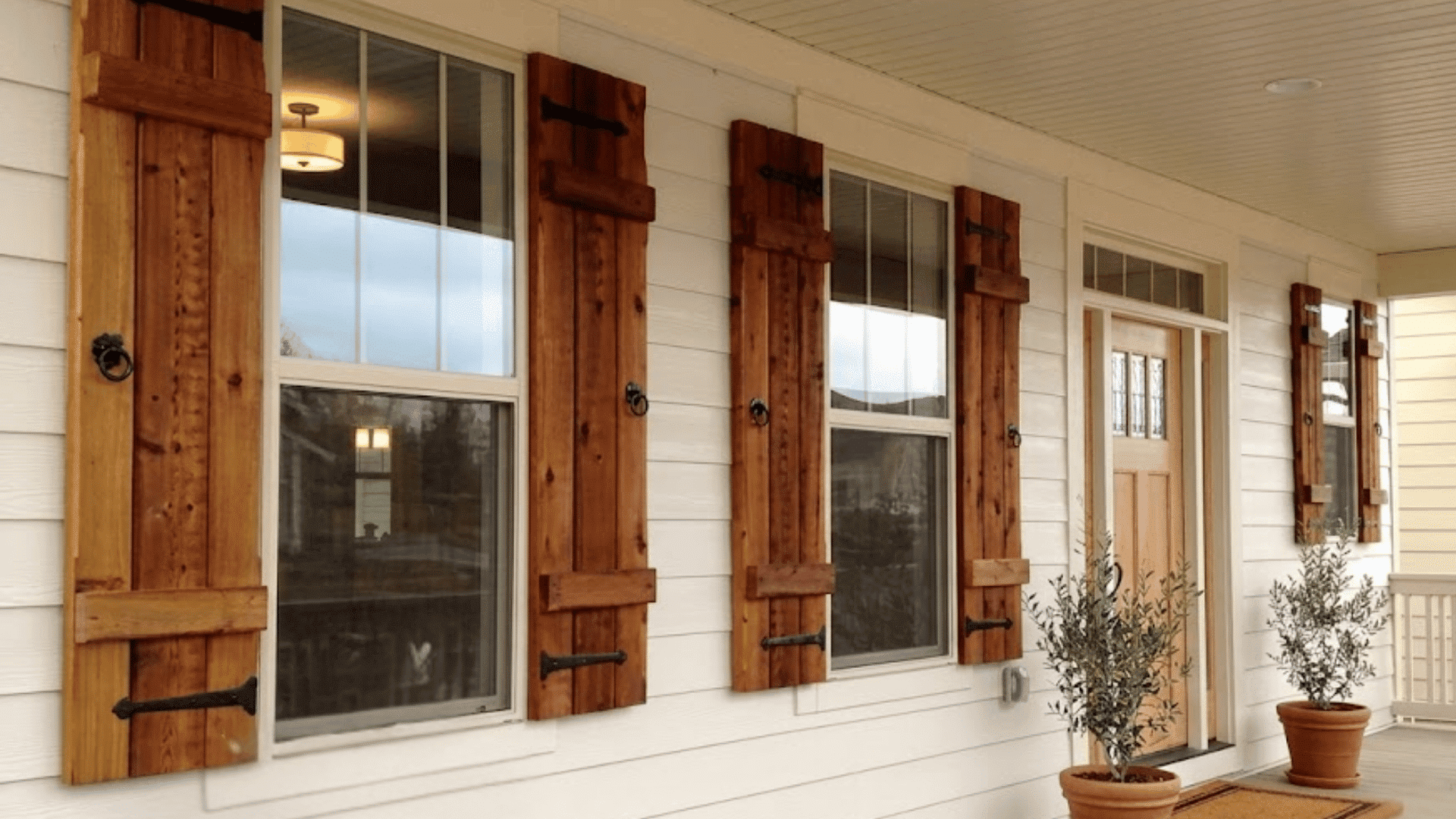 A long covered porch on a house has white siding wooden pillars and shutters plus potted olive trees