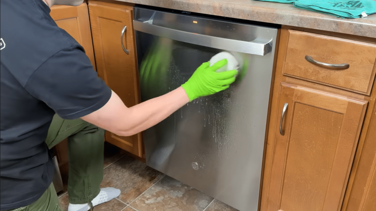 a person wearing a green glove scrubs the exterior stainless steel door of a dishwasher with a sponge