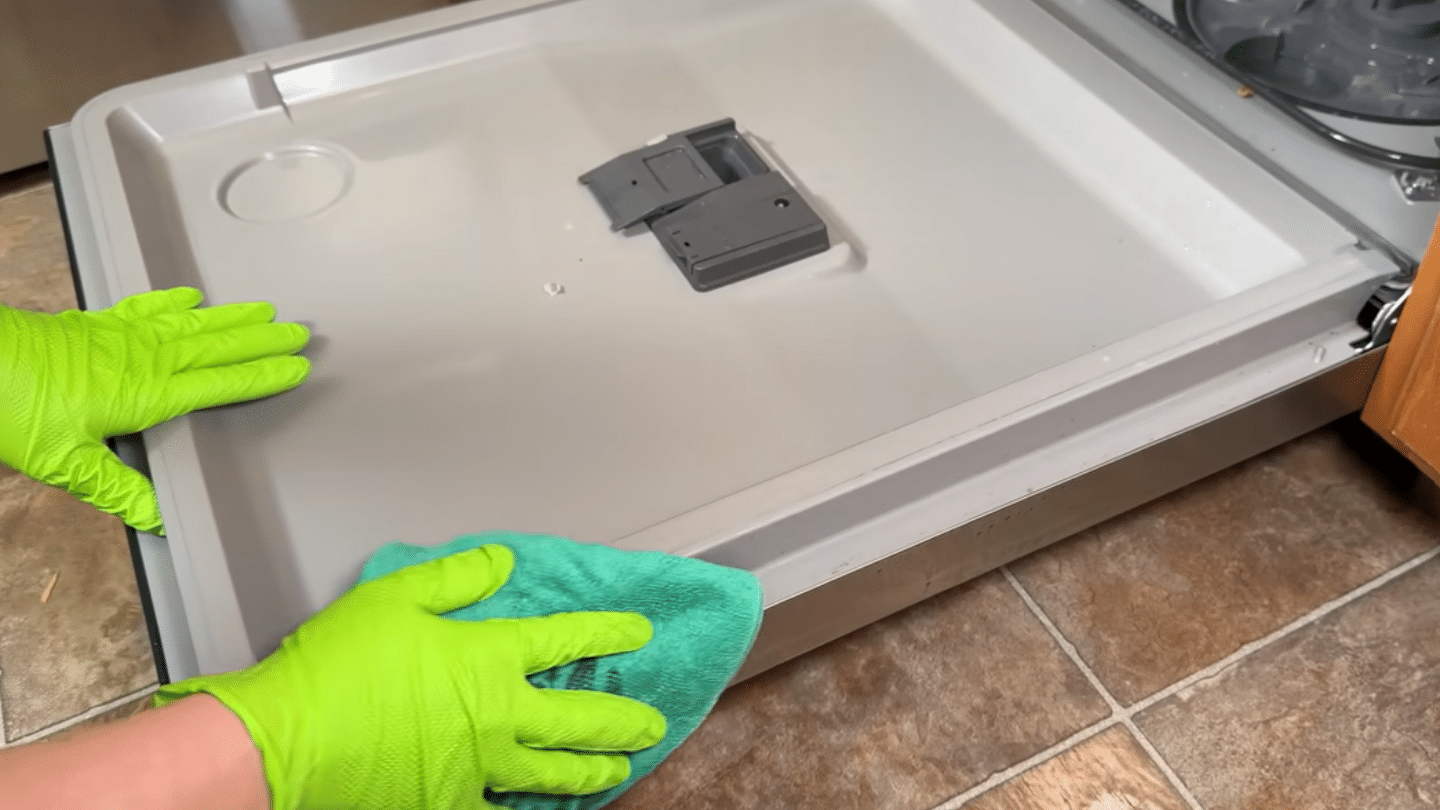 a person wearing bright green gloves uses a teal cloth to wipe down the inside edges of an open dishwasher door