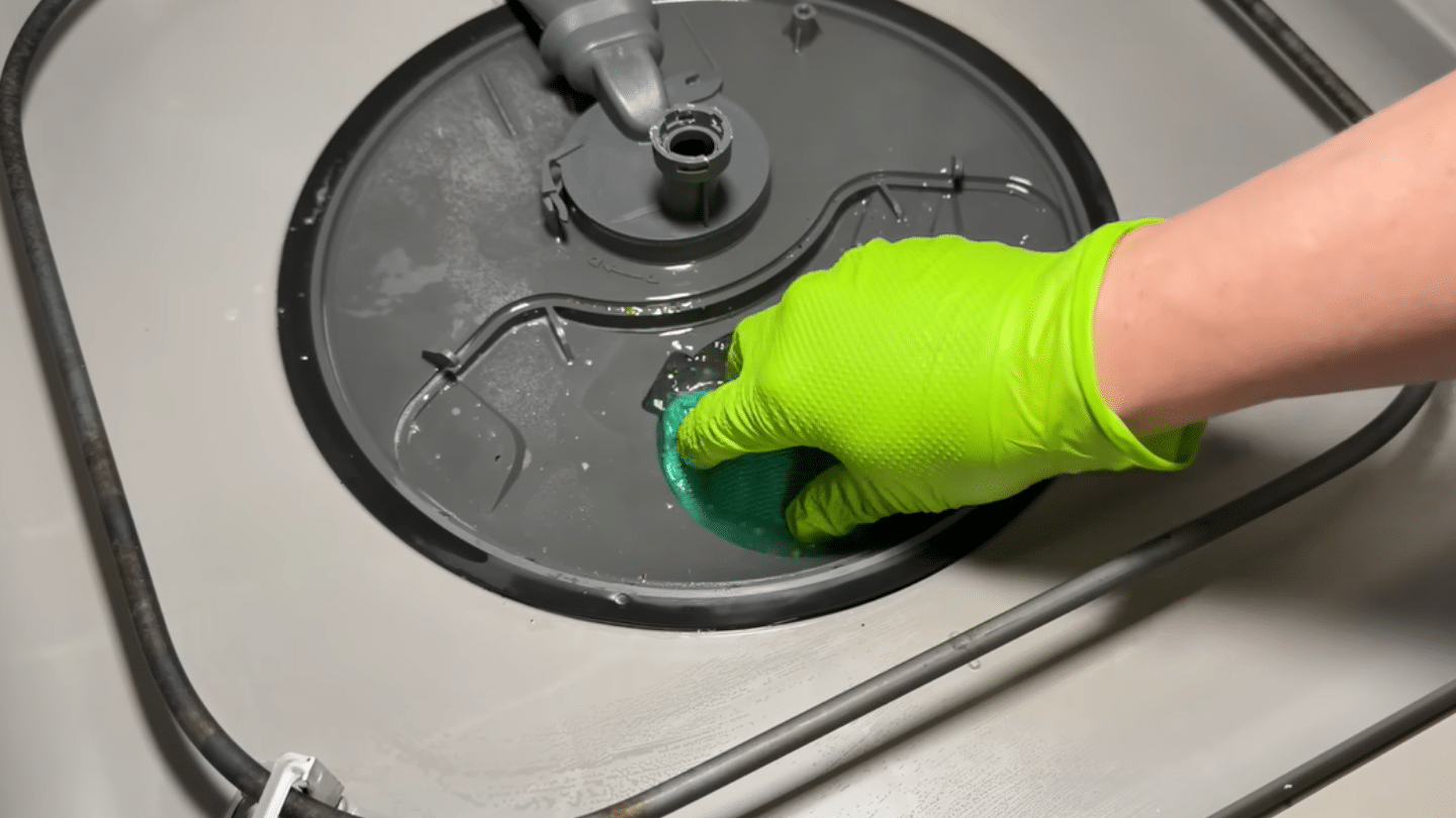 a person wearing a bright green glove using a sponge to scrub the inside base of a grey dishwasher