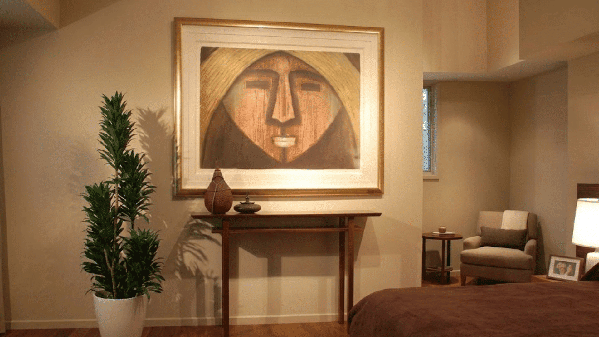 bedroom corner with framed abstract portrait above a wooden console table beside a tall potted plant and armchair seating area