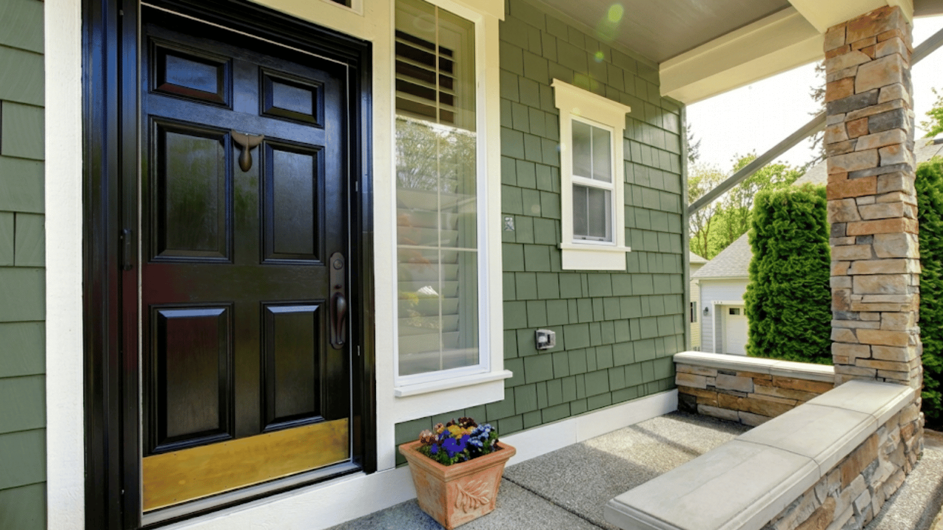 black front door with white door casing beside green siding and stone porch columns with flower planter