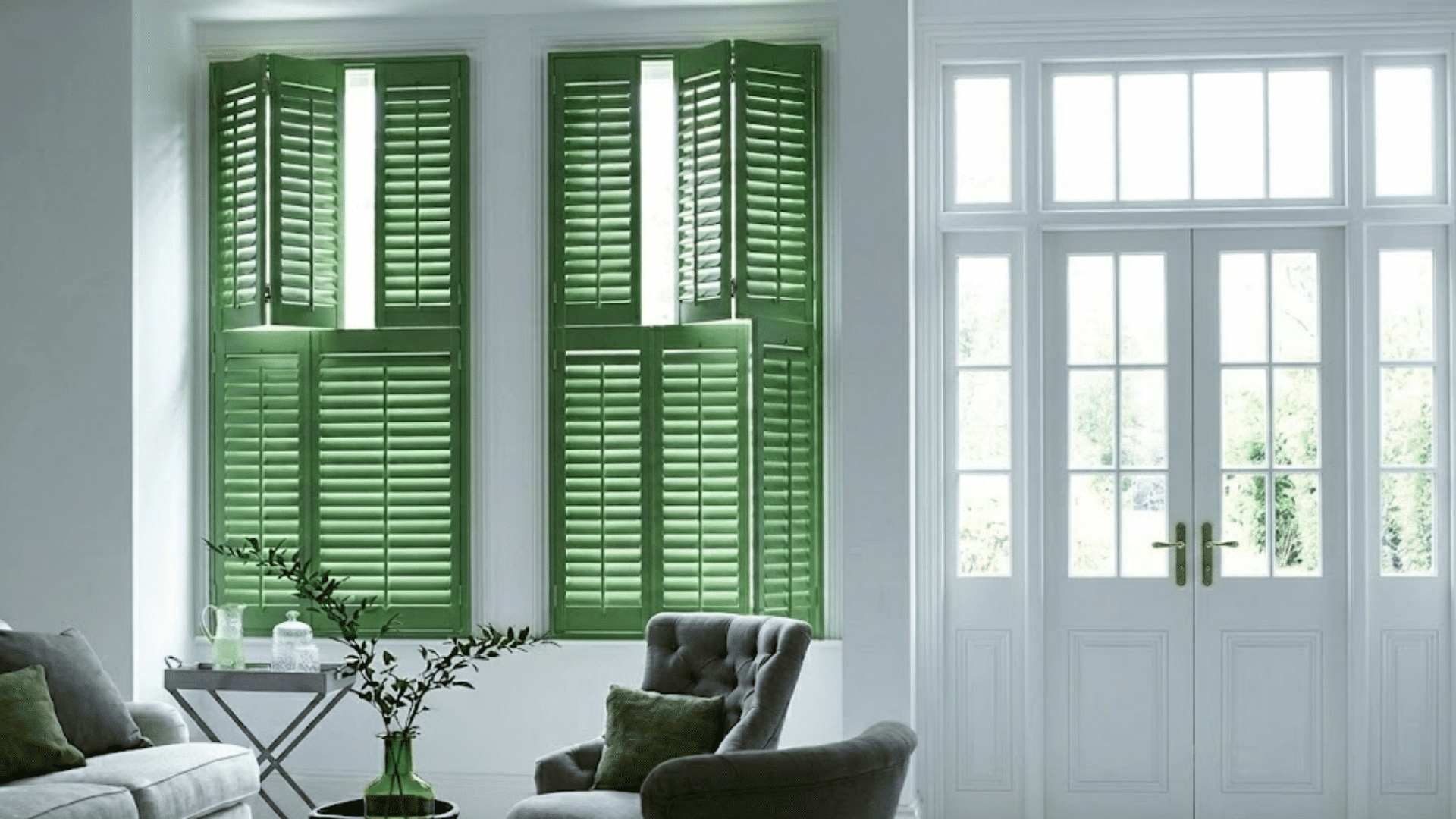 bright living room with green shuttered windows white french doors and cushioned chairs beside a sofa and vase