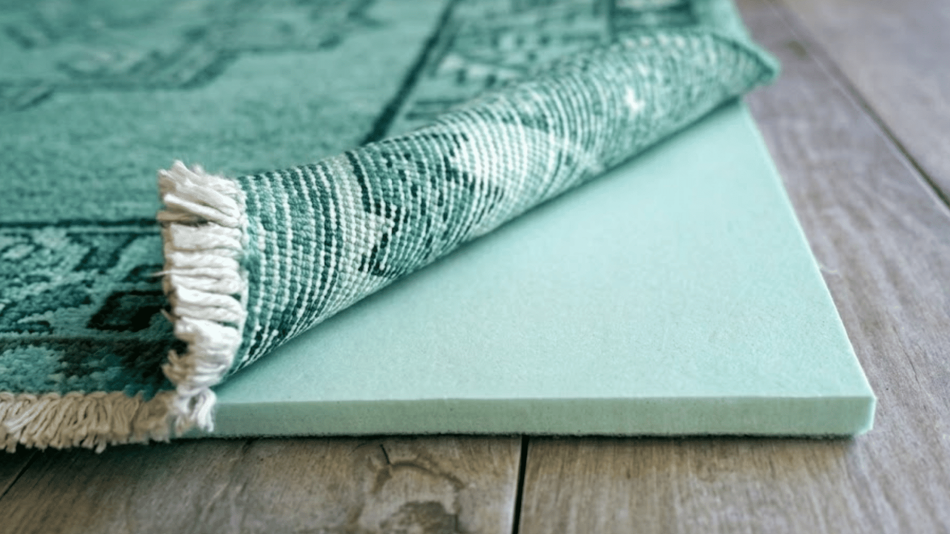 corner of a teal patterned area rug lifted to reveal a foam underlay cushion placed on a hardwood floor surface