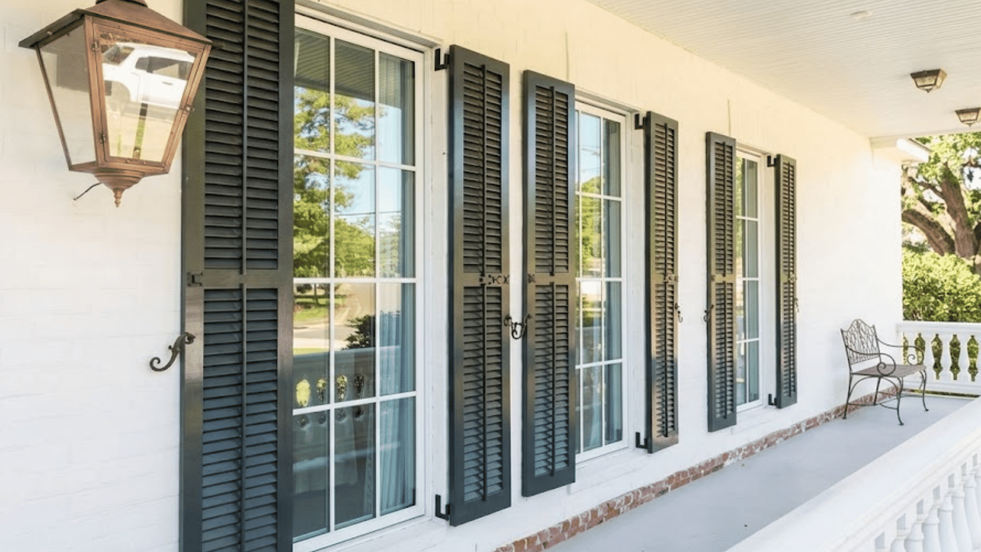 covered porch with black shutters framing tall windows beside a metal chair and white railing overlooking trees