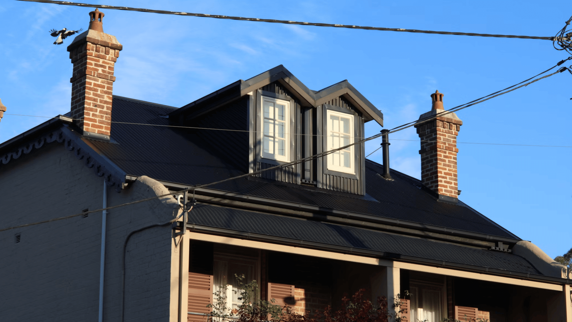 dark metal roof with dormer windows brick chimneys and overhead utility wires against blue sky