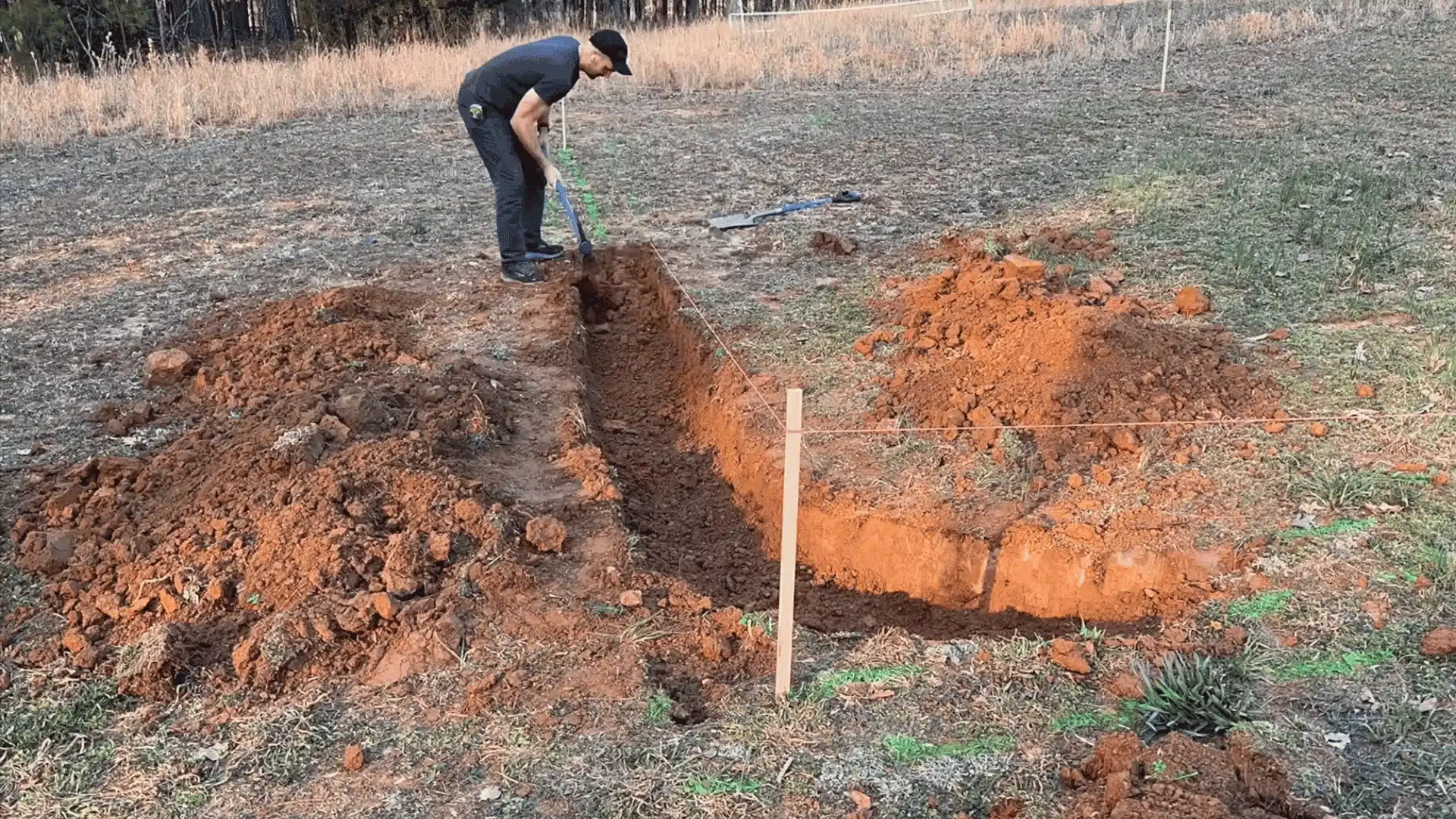 Man digging a trench in a grassy field using a shovel, with marked string lines and stakes outlining layout near soil piles.