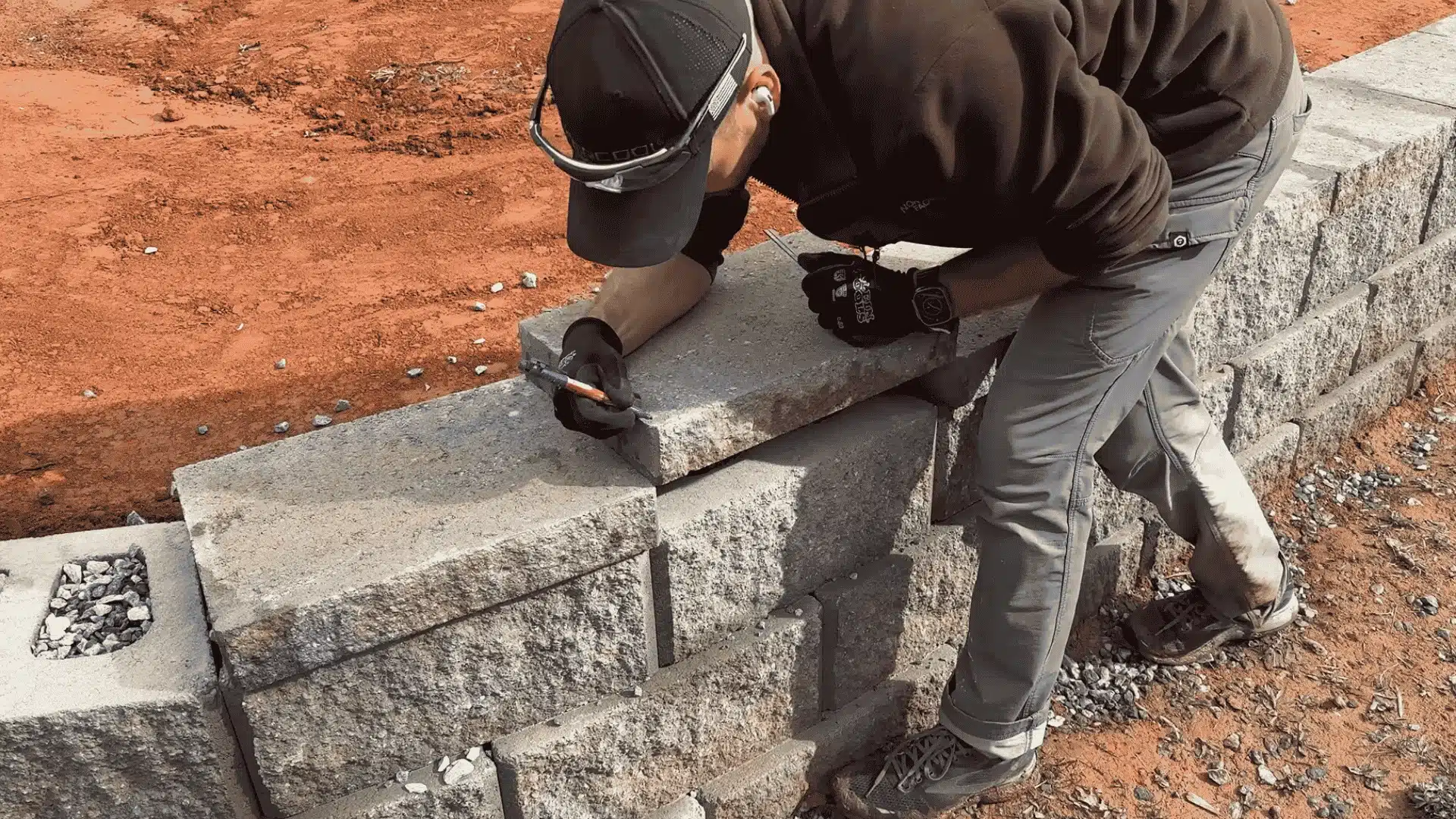 Worker marking and cutting a stone retaining wall block while kneeling on a partially built wall with red soil background.